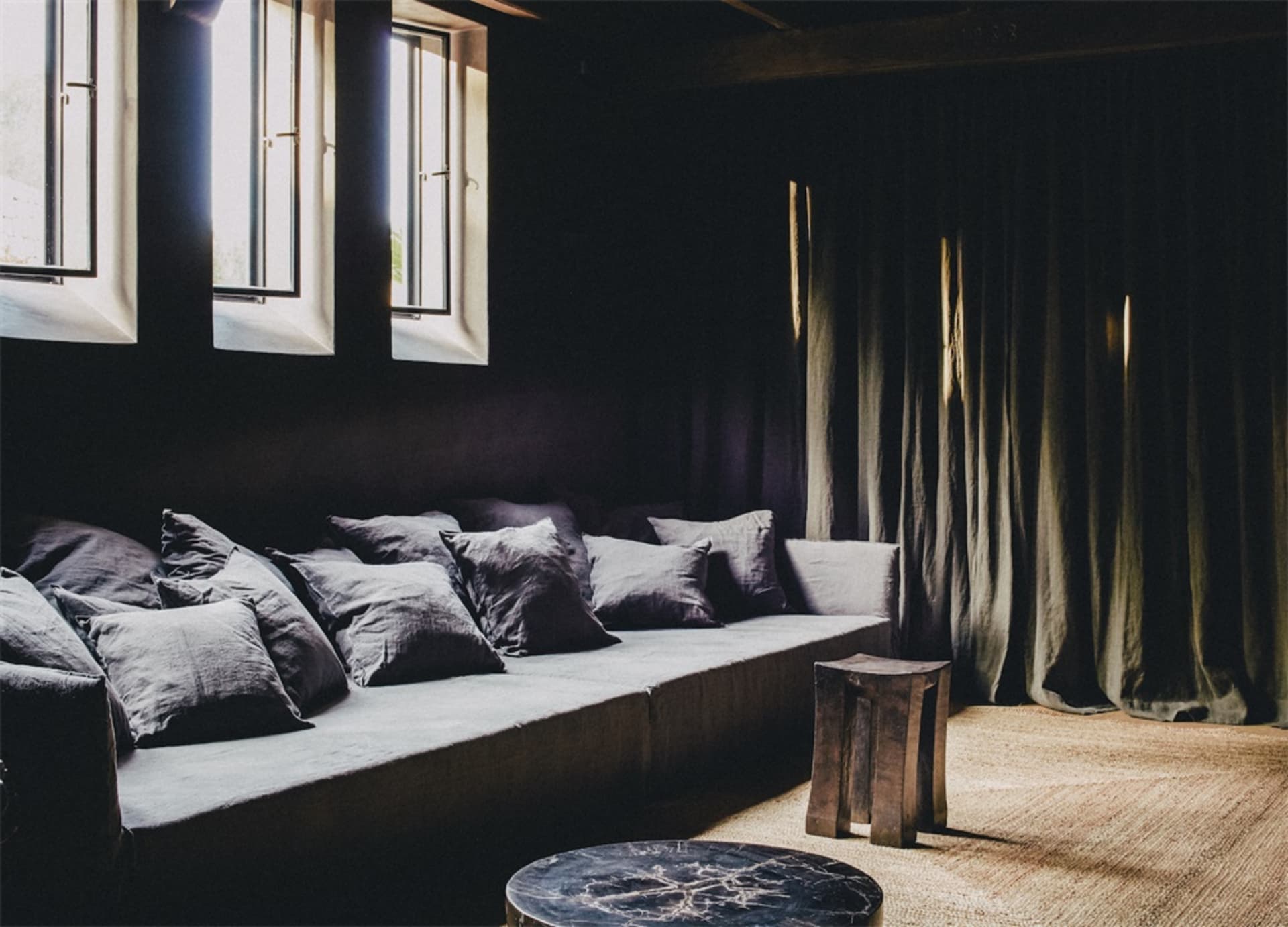 Industrial-style living room with a long gray couch adorned with multiple fabric cushions, large windows with curtains, and a
