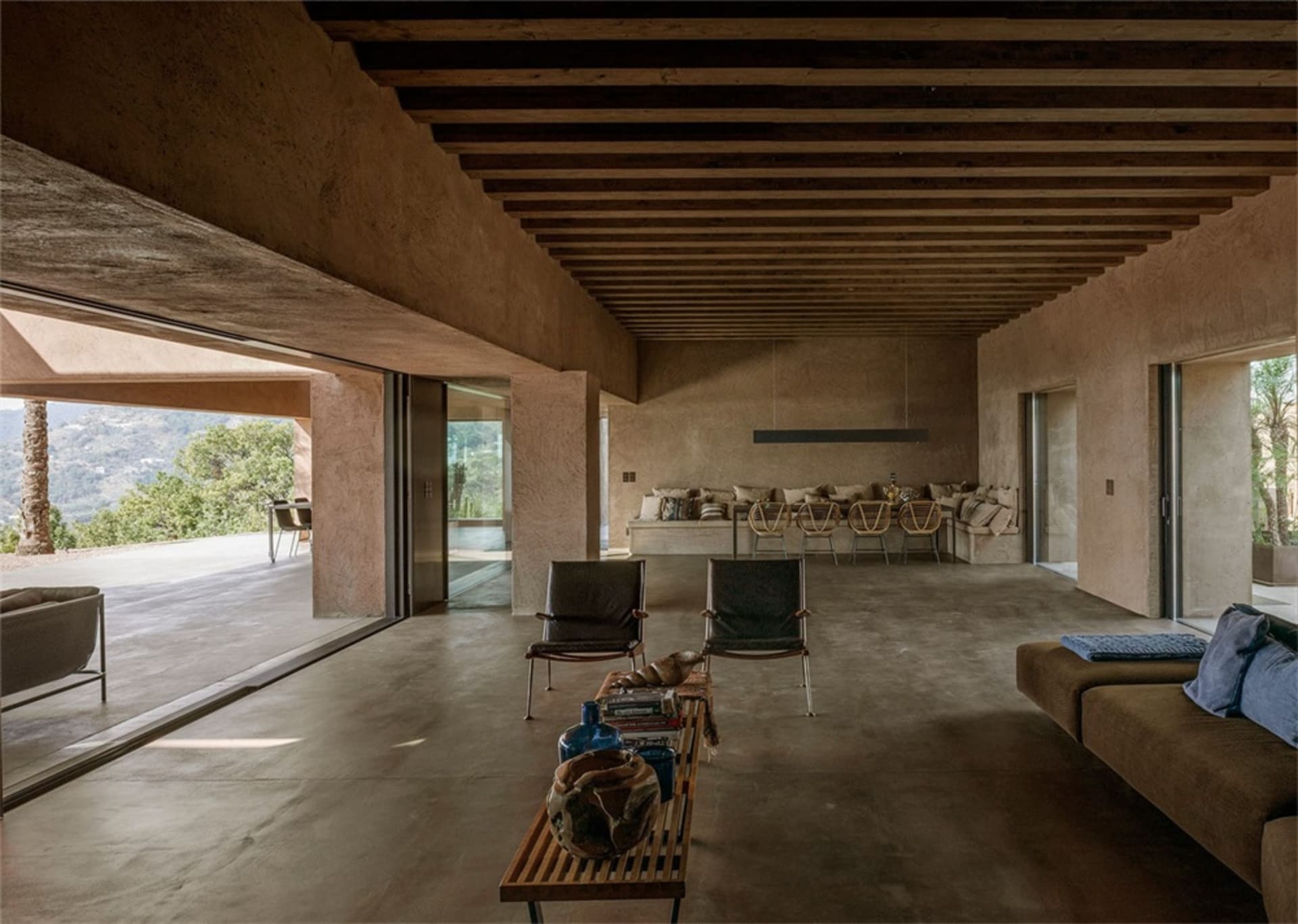 A modern living room with concrete flooring, minimalist furniture, and large windows providing a view of the outdoor area.