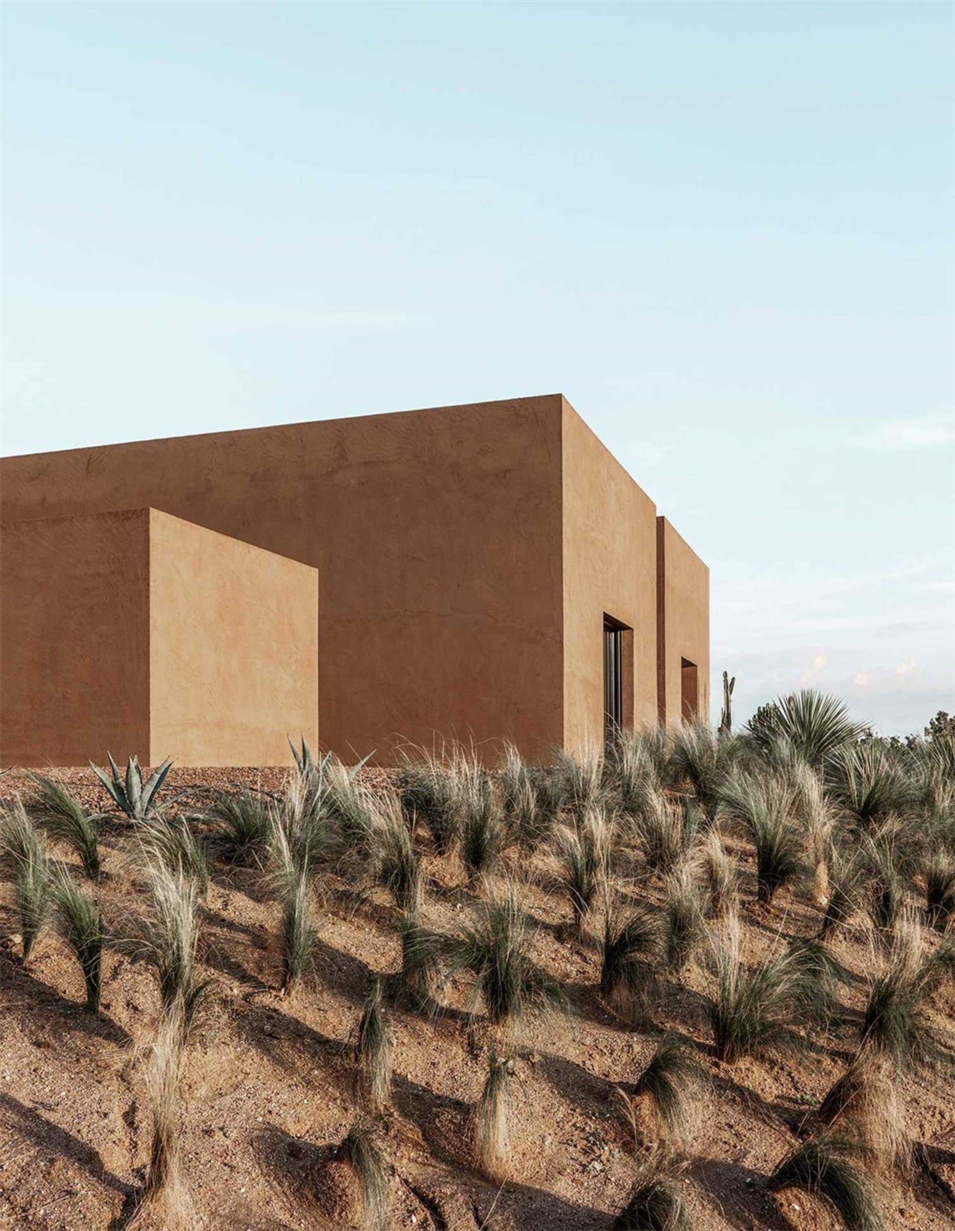 A modern minimalist house with a clay stucco exterior surrounded by desert vegetation.