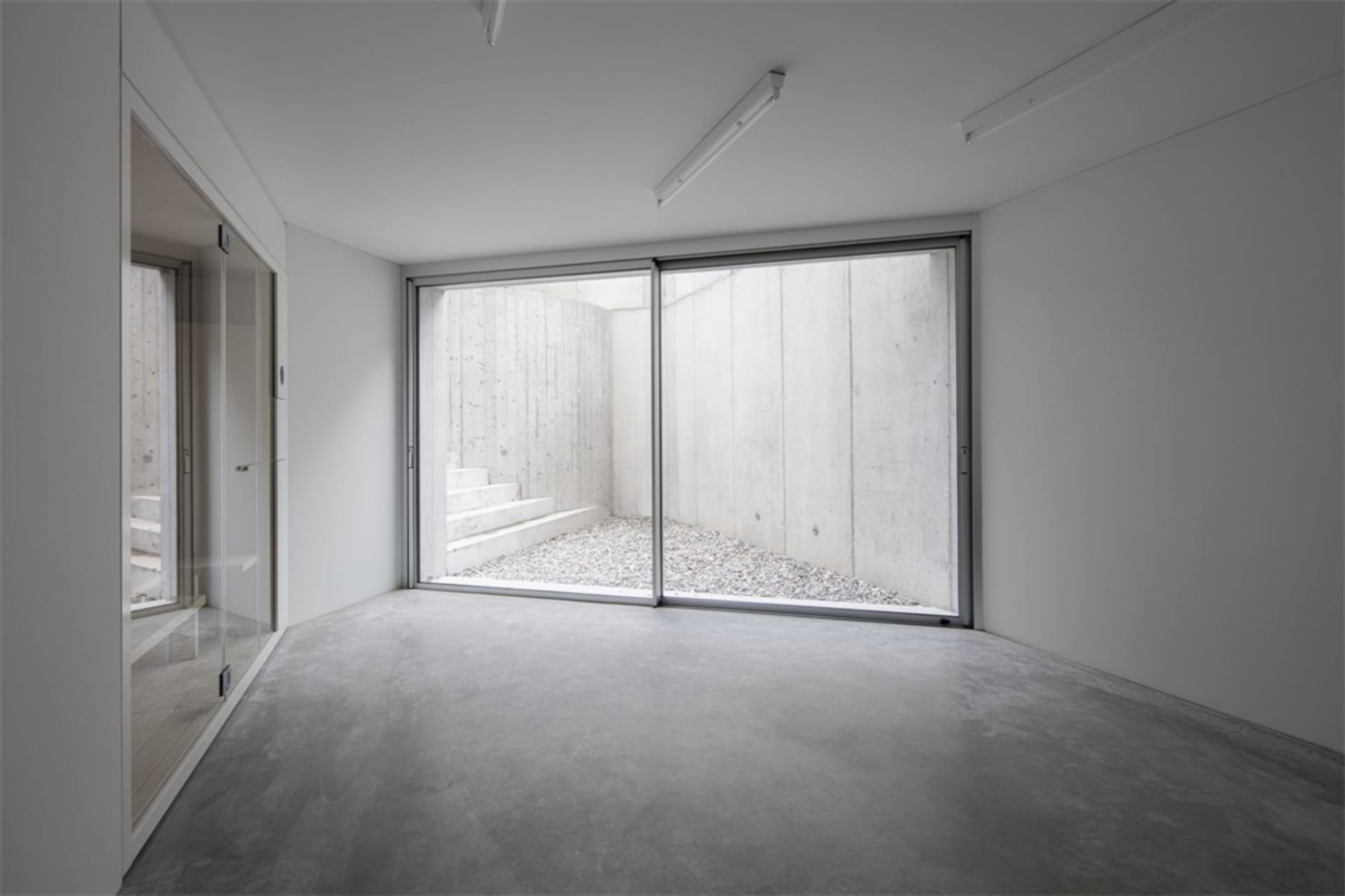 A minimalist space with a concrete ground and wall, featuring a view of a gravel courtyard and steps leading downwards.