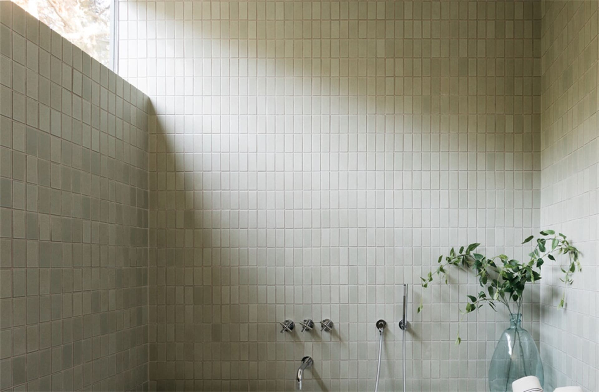 A modern bathroom with white tiled walls and floor, featuring a vase with green leaves and a window letting in natural light.
