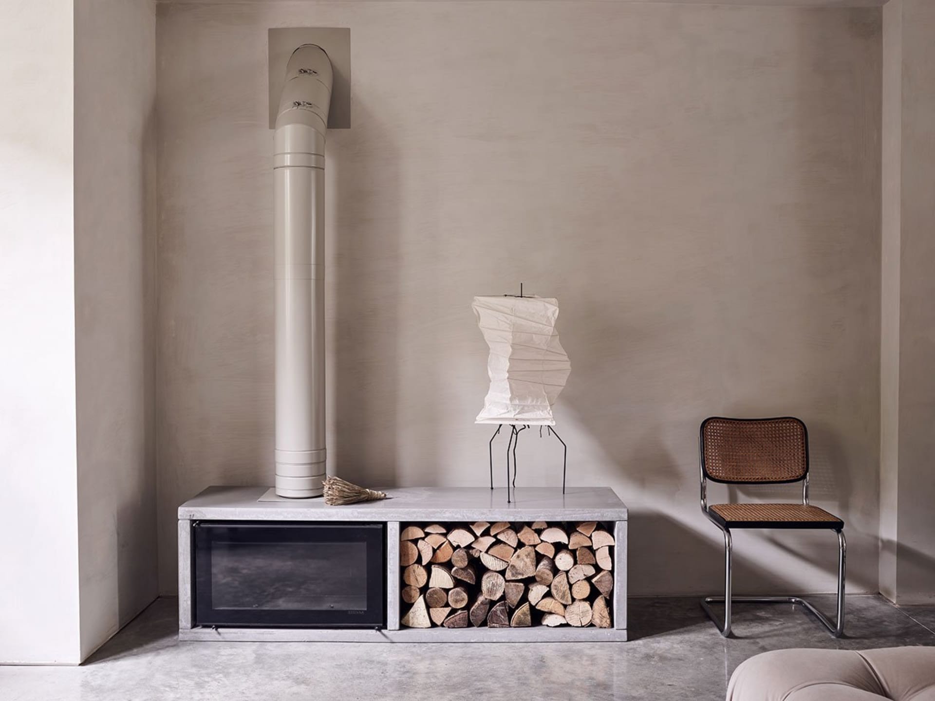 A minimalist living room with a concrete fireplace and wood storage beneath it, featuring a concrete floor and a modern chair