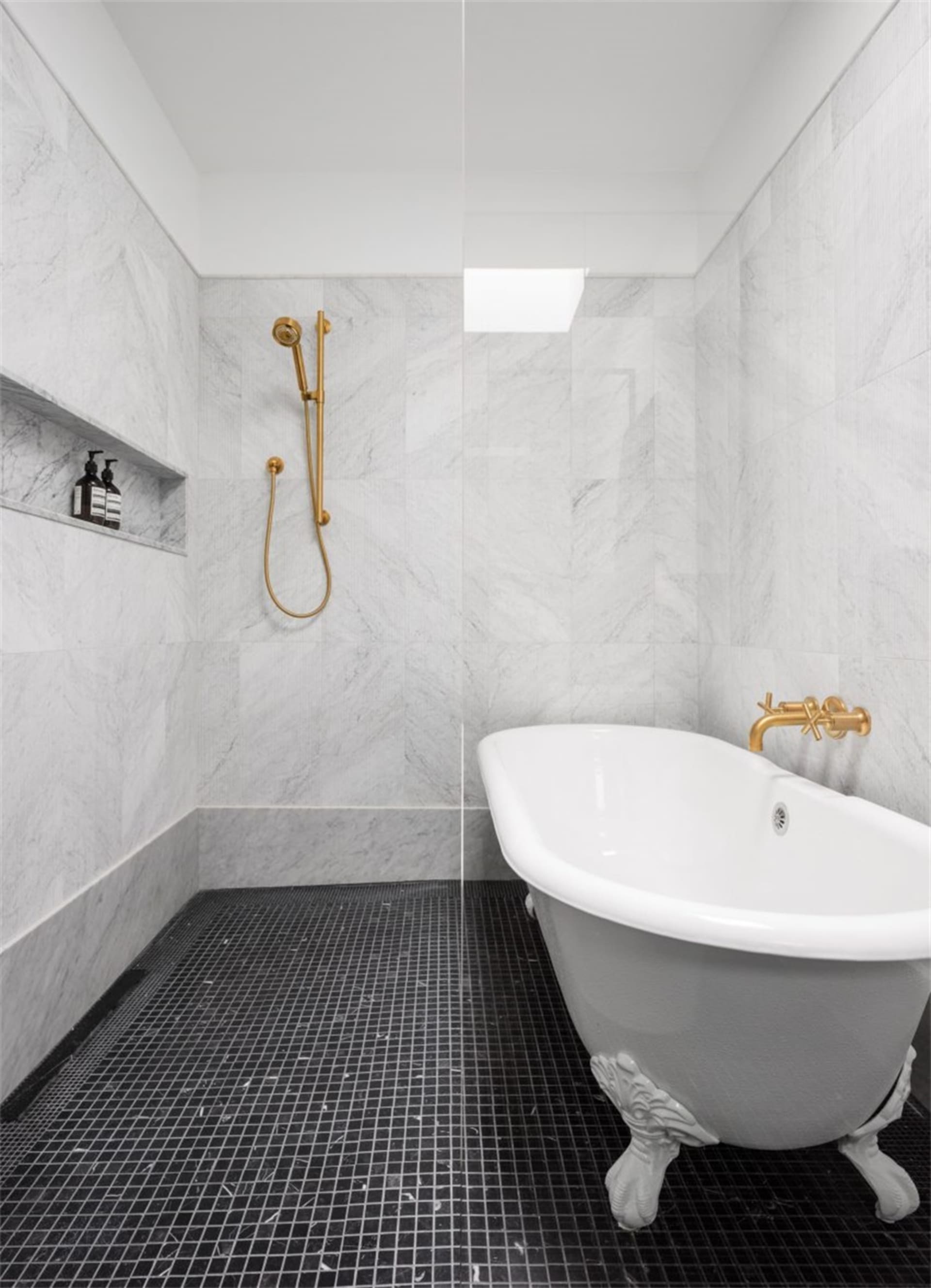 A minimalist bathroom with marble walls and porcelain tiles, featuring a clawfoot bathtub and a walk-in shower with gold fixt