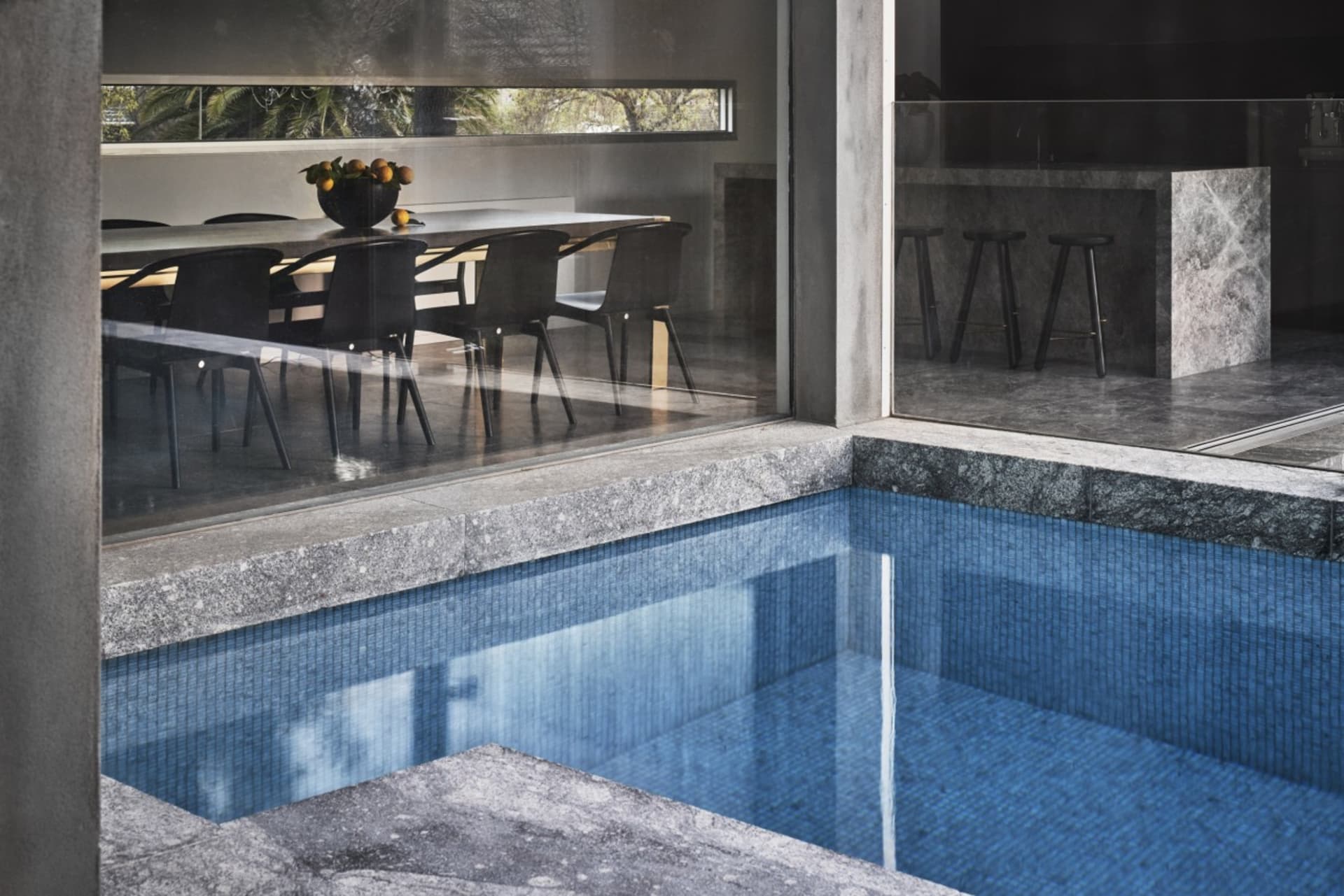 A modern indoor swimming pool with granite tiles, featuring a table with oranges in the background.