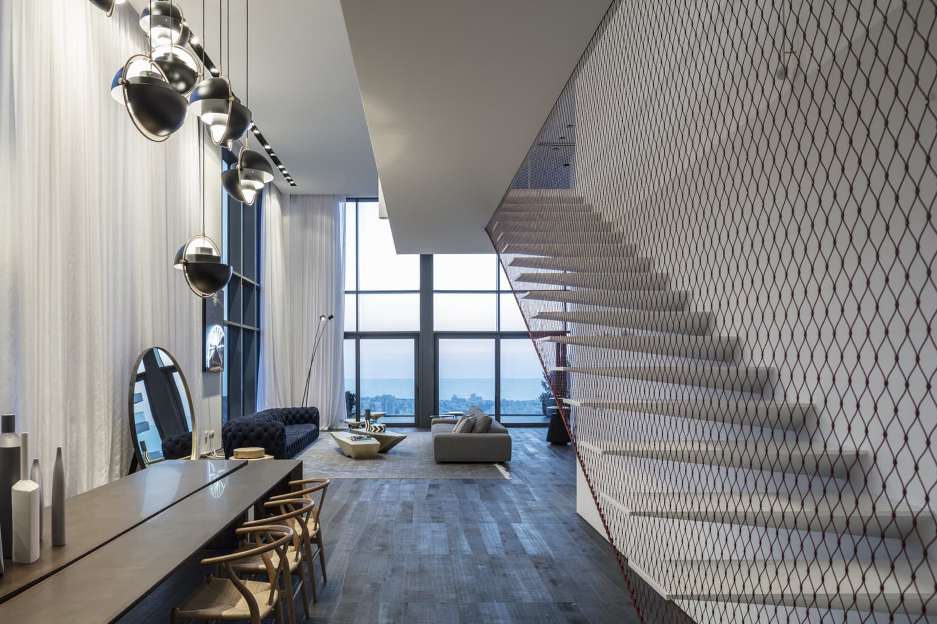 A modern living room with wooden accents, featuring a large table and chairs, a couch, and a staircase with a net railing.