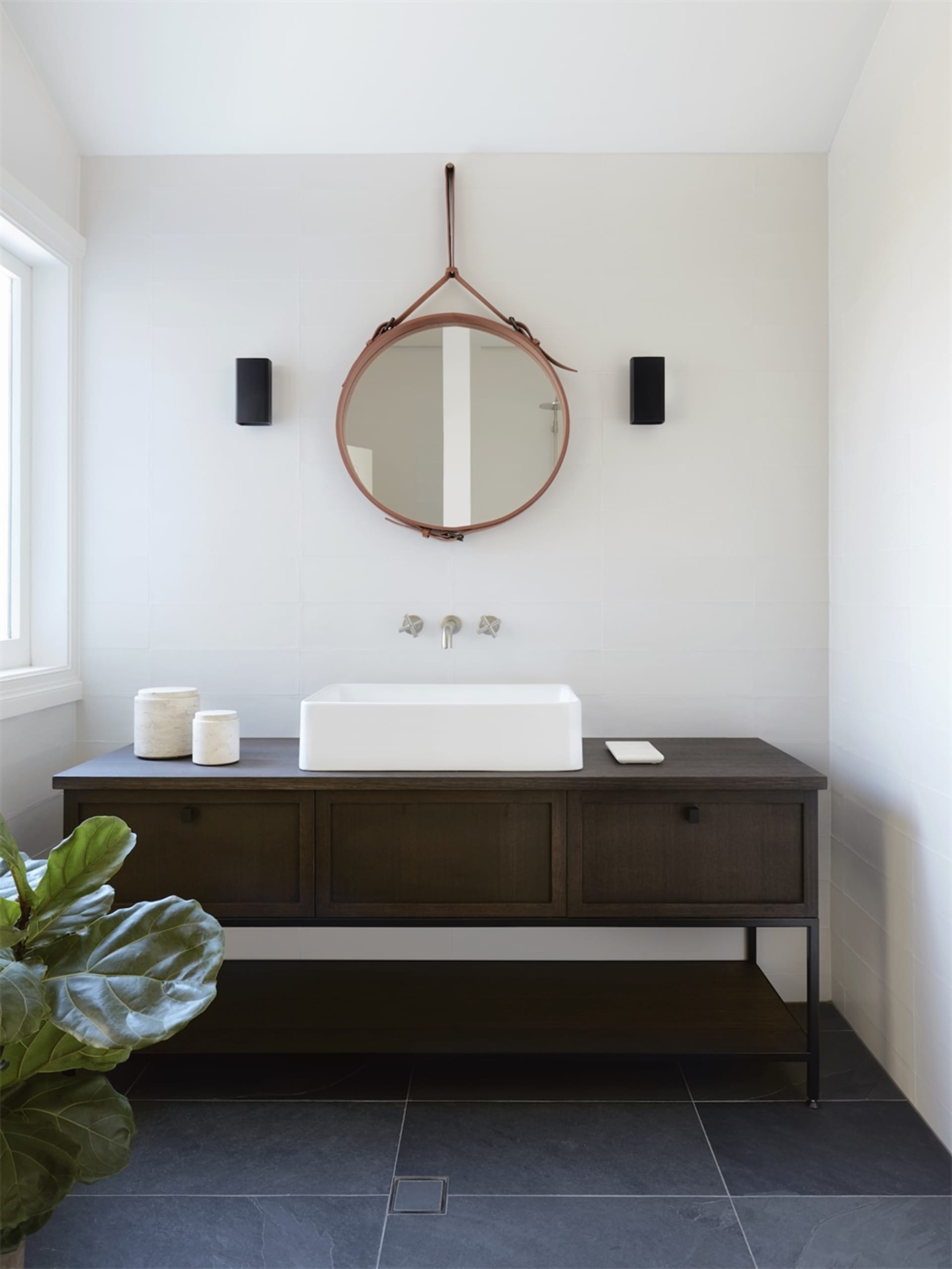 Modern minimalist bathroom featuring a dark wooden vanity with a ceramic sink basin, a circular leather-framed mirror, and wh