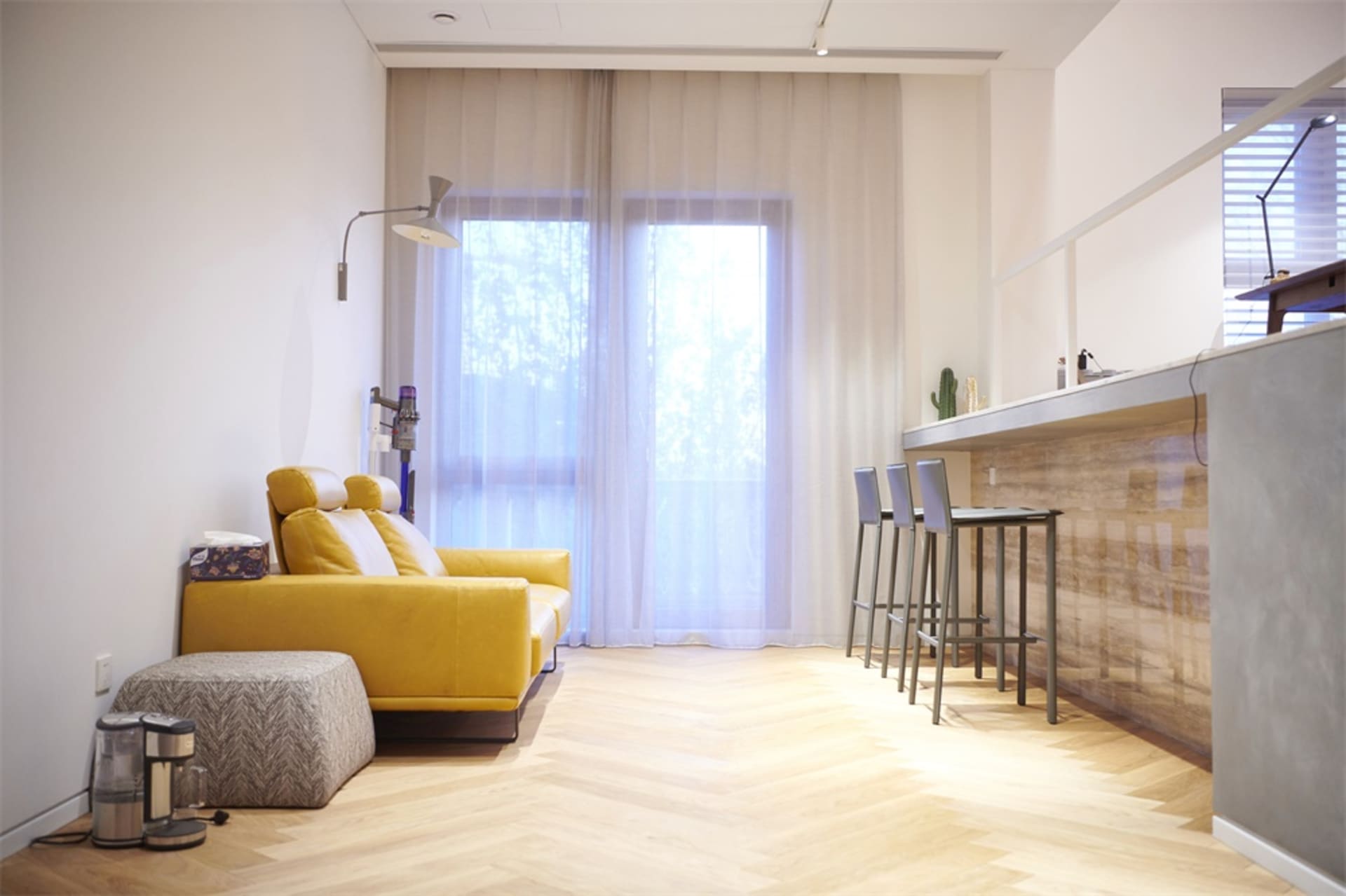 A spacious, modern living room with a yellow sofa, hardwood floors, and sheer curtains.