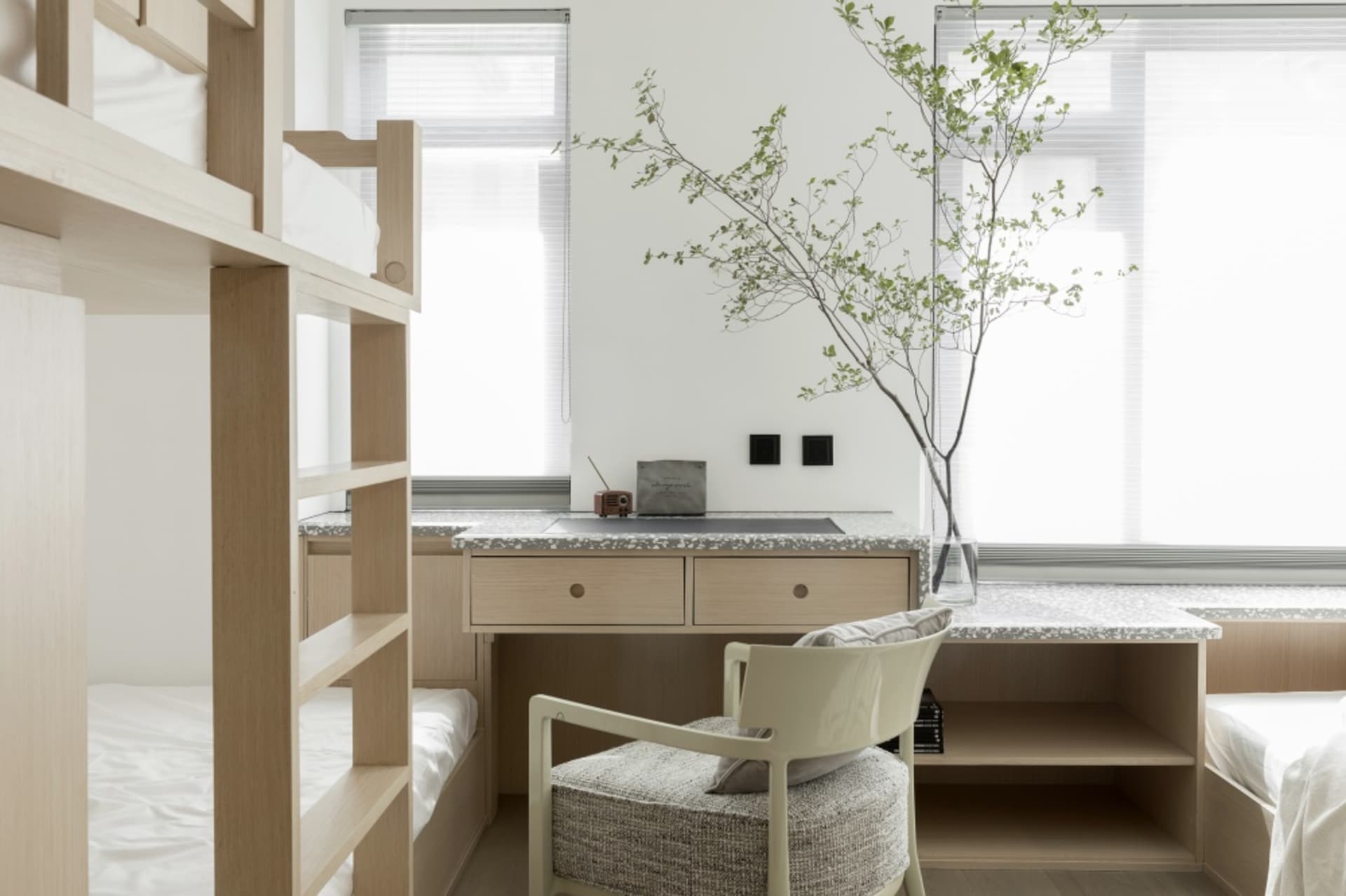 A minimalist modern bedroom interior featuring a wooden bunk bed and a stone-topped desk with a plant on it.
