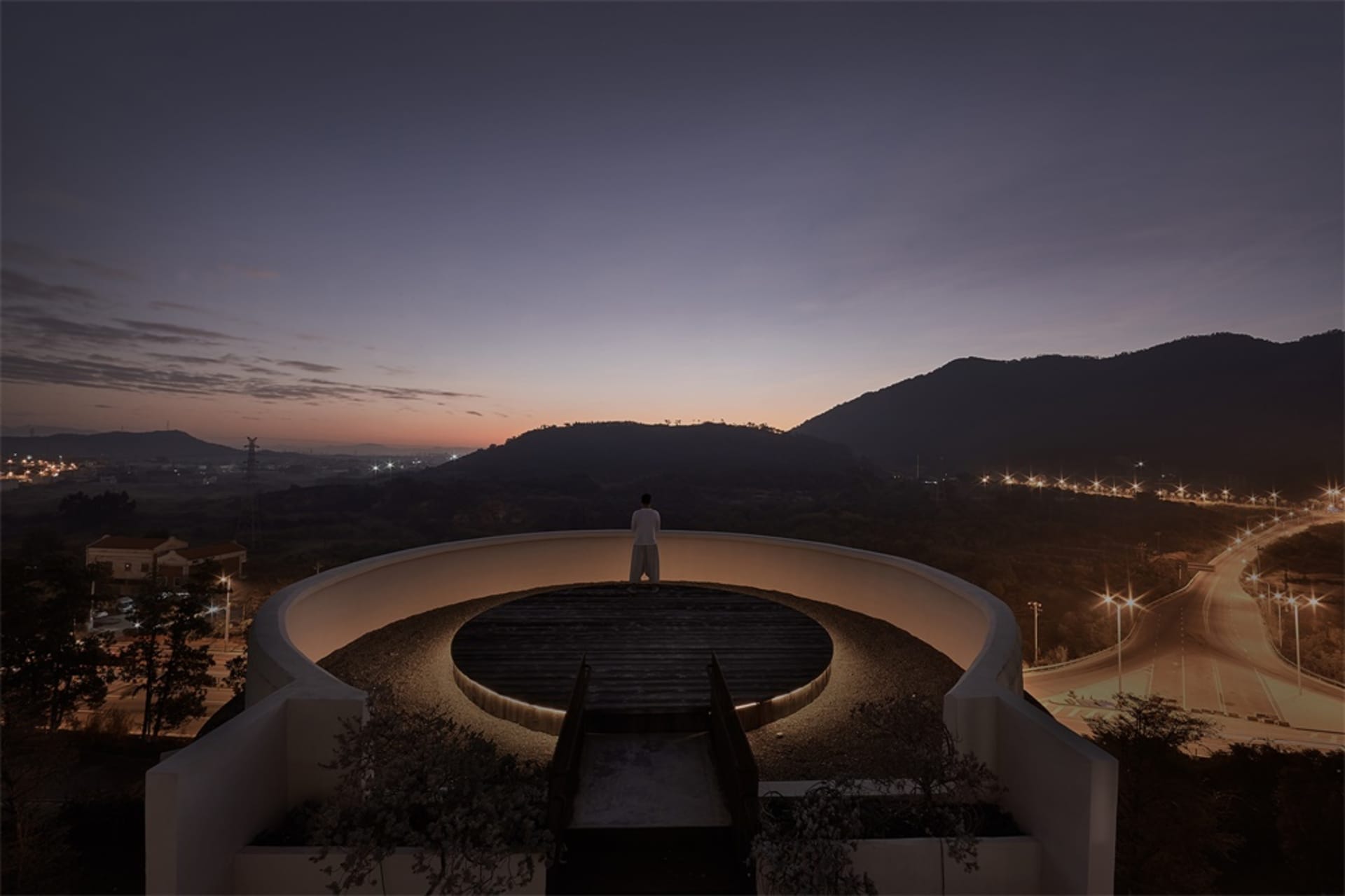 A person standing on a circular terrace overlooking a valley at sunset.