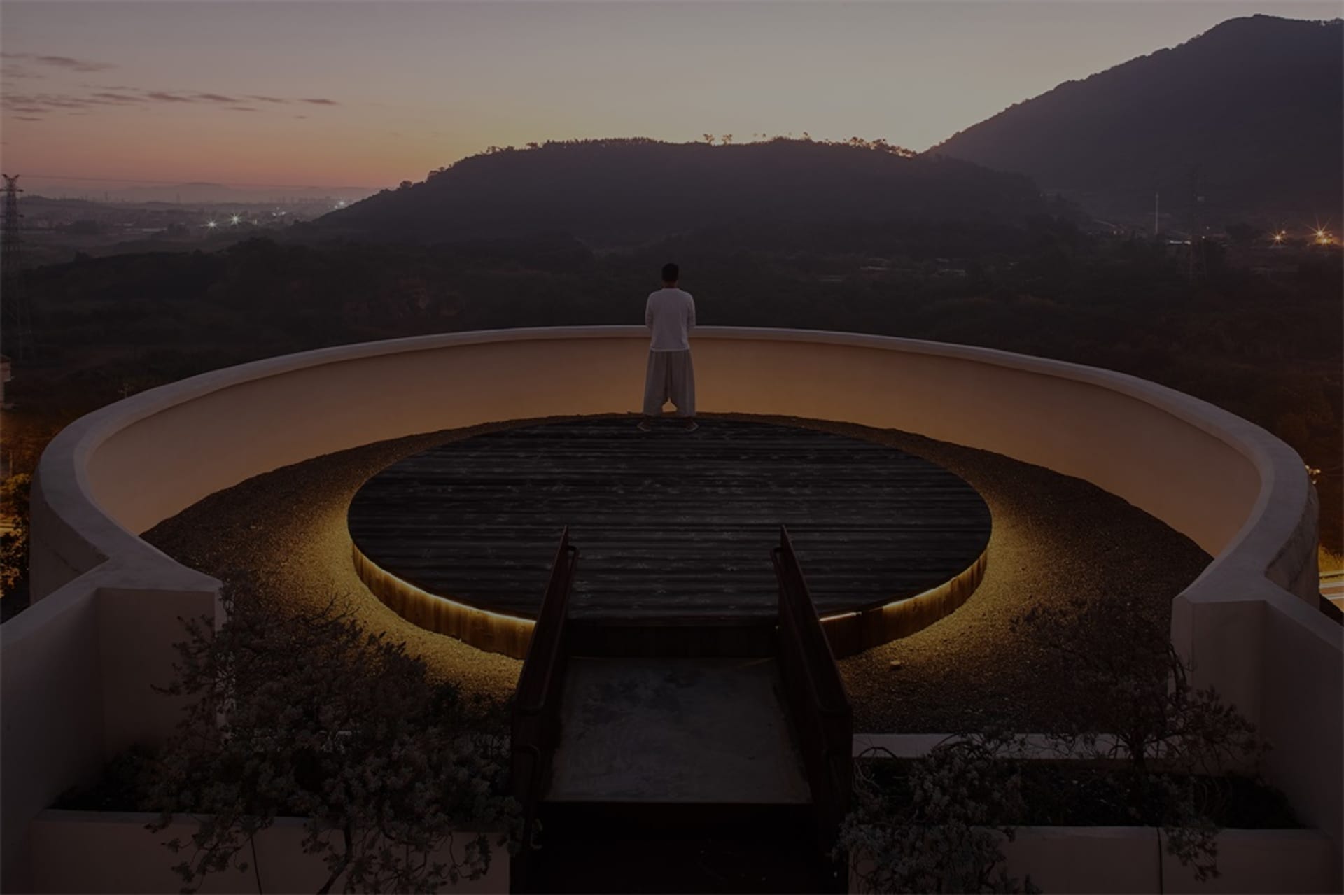 Person standing on a circular wooden platform with illuminated edges, overlooking a mountainous landscape at sunset.