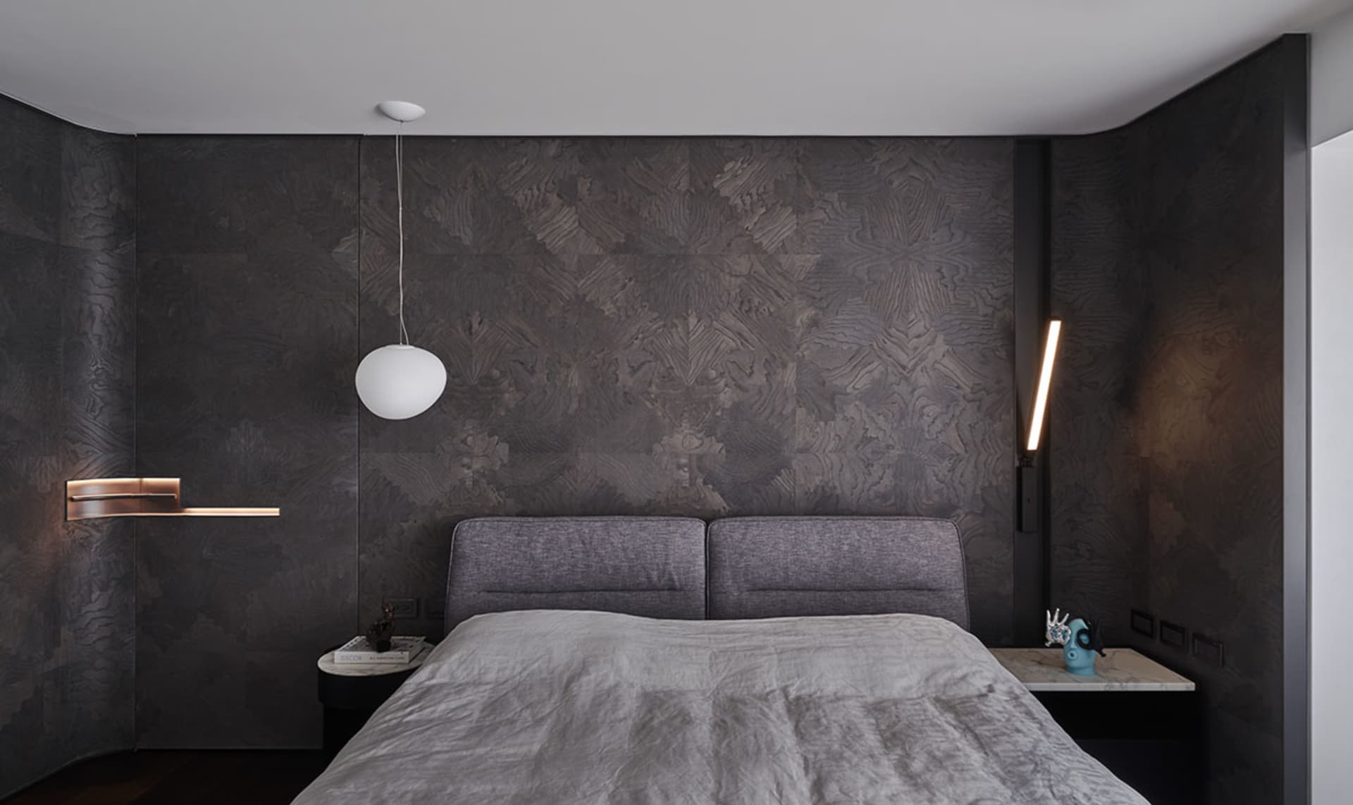 A modern bedroom featuring a textured dark wall, a grey upholstered bed, and minimalistic design elements.