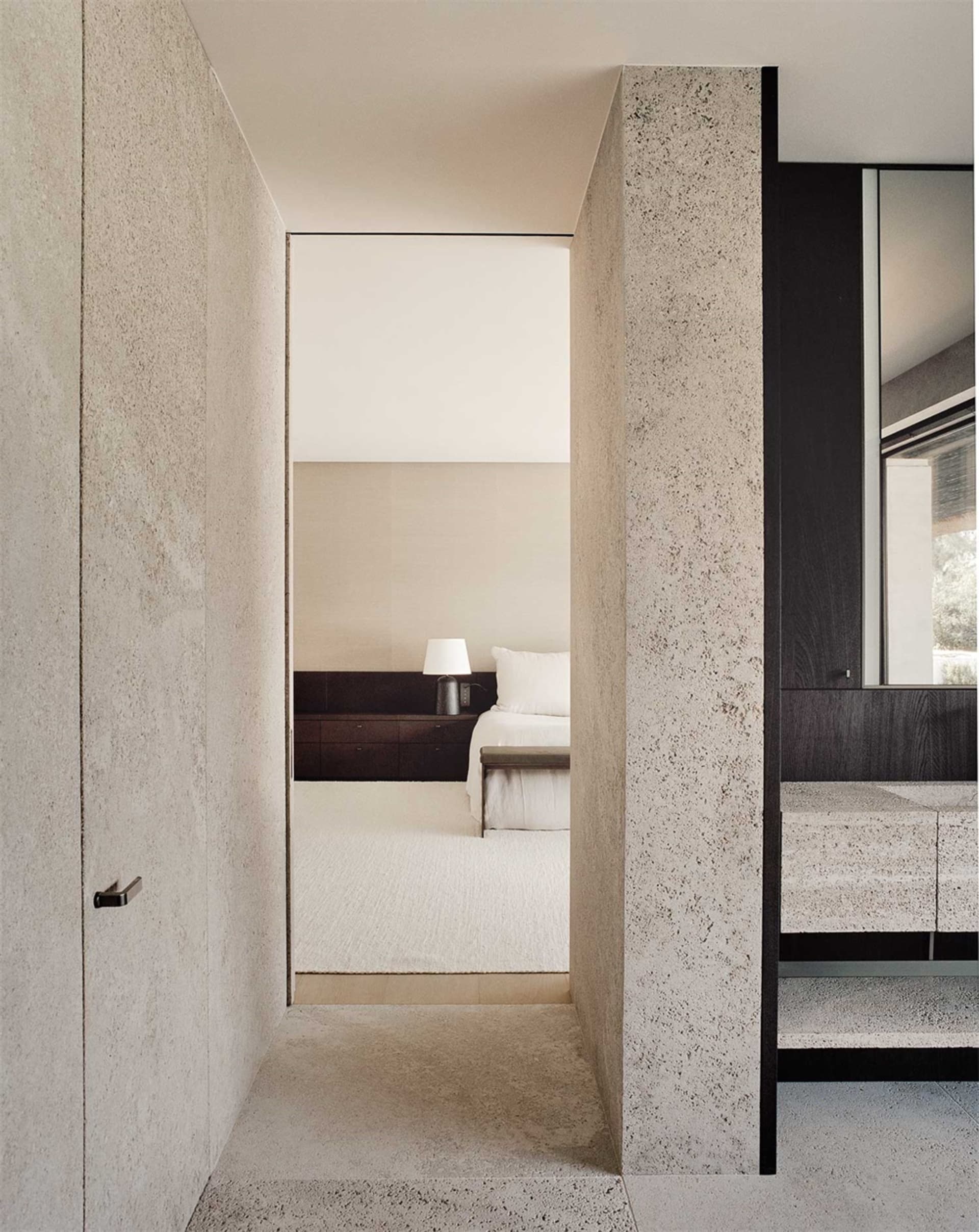 Minimalist bedroom with concrete walls and floor, featuring a bed with white sheets and pillows, and a nightstand with a lamp