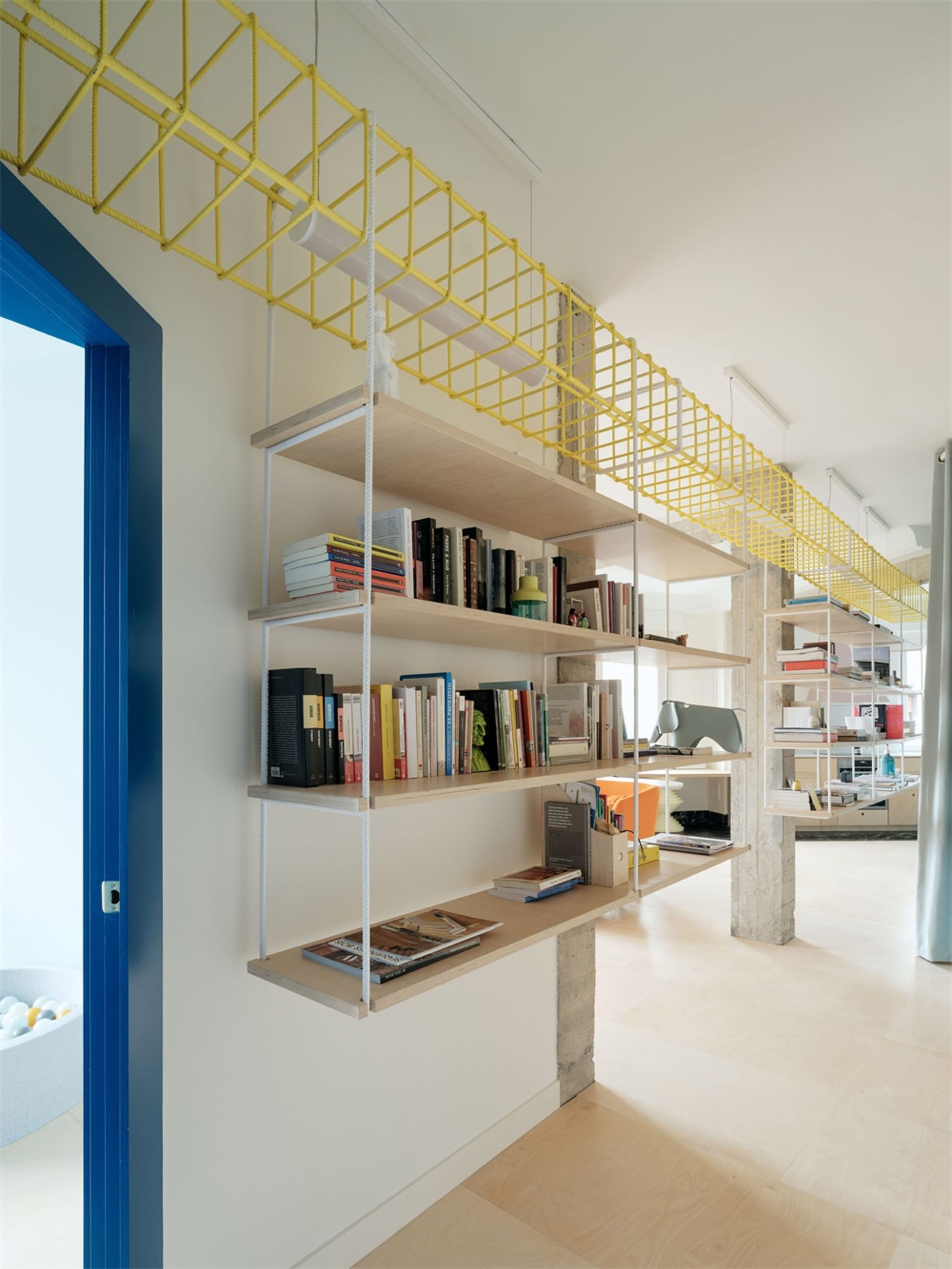 A modern interior with wooden shelves displaying books and other items, connected to the ceiling by a yellow rope structure.