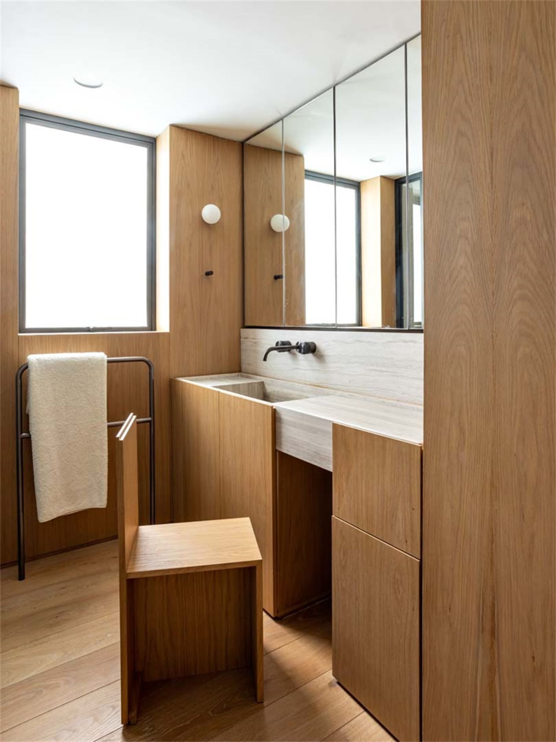 Modern bathroom with minimalist wooden cabinets, sink, and stool, featuring a large mirror and natural light through the wind