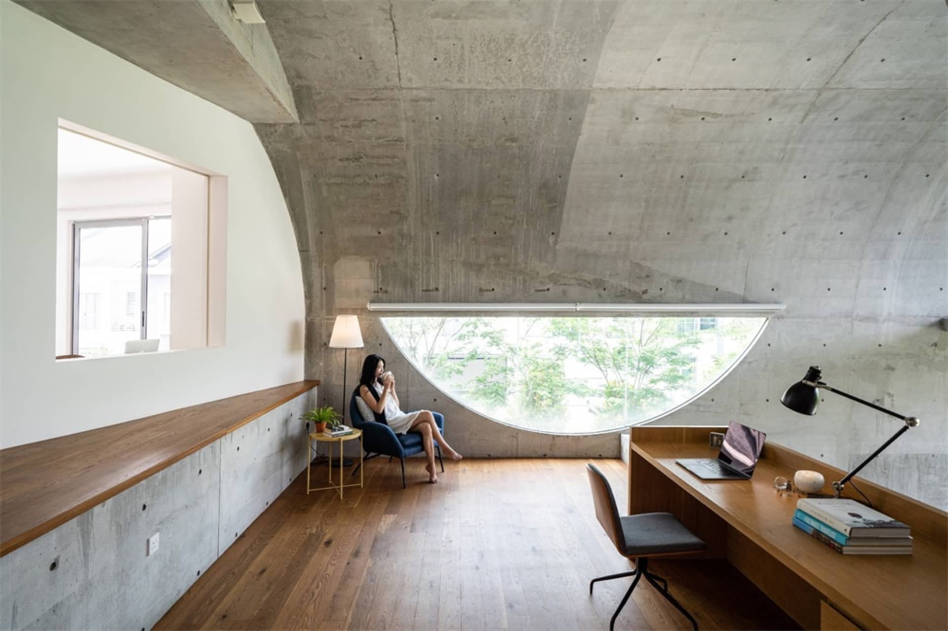A modern home office features a concrete console with a built-in desk, a woman sitting on a blue armchair by a round window,