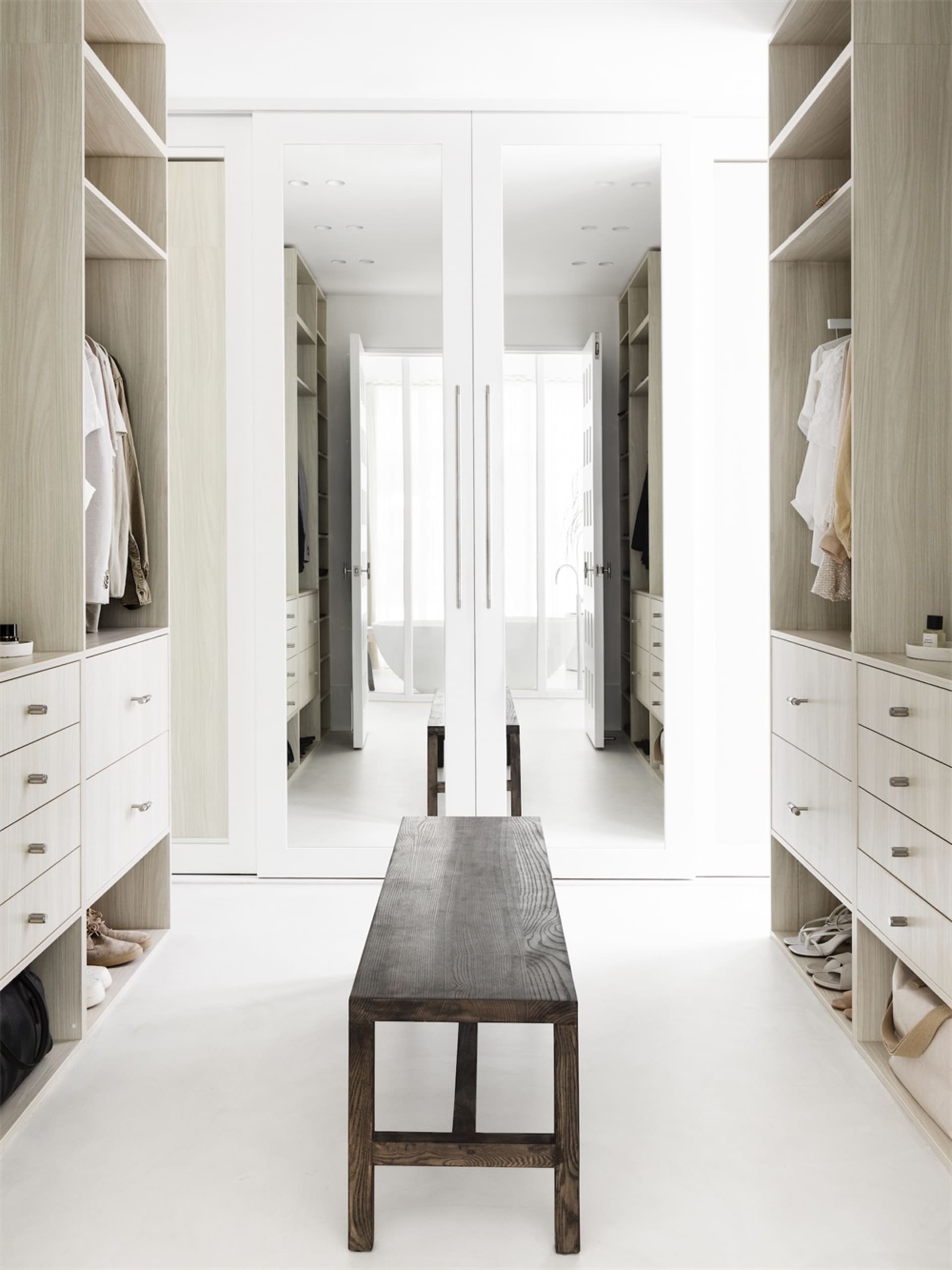 A modern white closet with open shelves, a wooden bench, and two large mirrors.