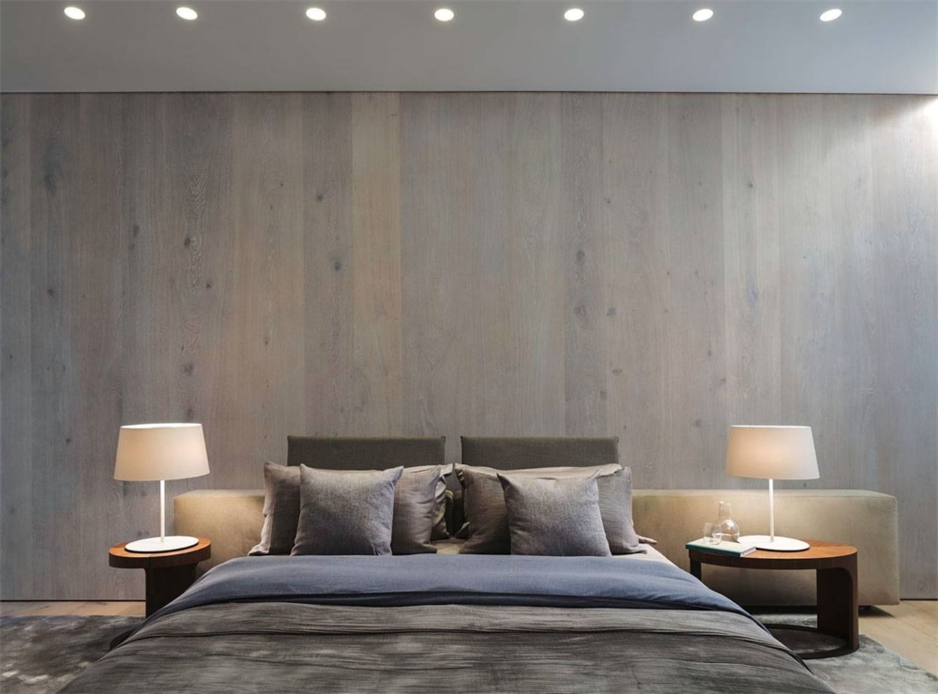 A minimalist bedroom with a wooden texture wall, grey bedding, and matching bedside tables with lamps.