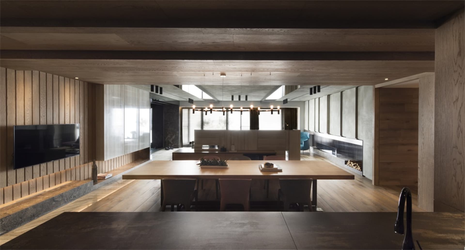 Modern minimalist living room featuring dark wooden flooring, walls, and furniture, with a large table and chairs, and a warm