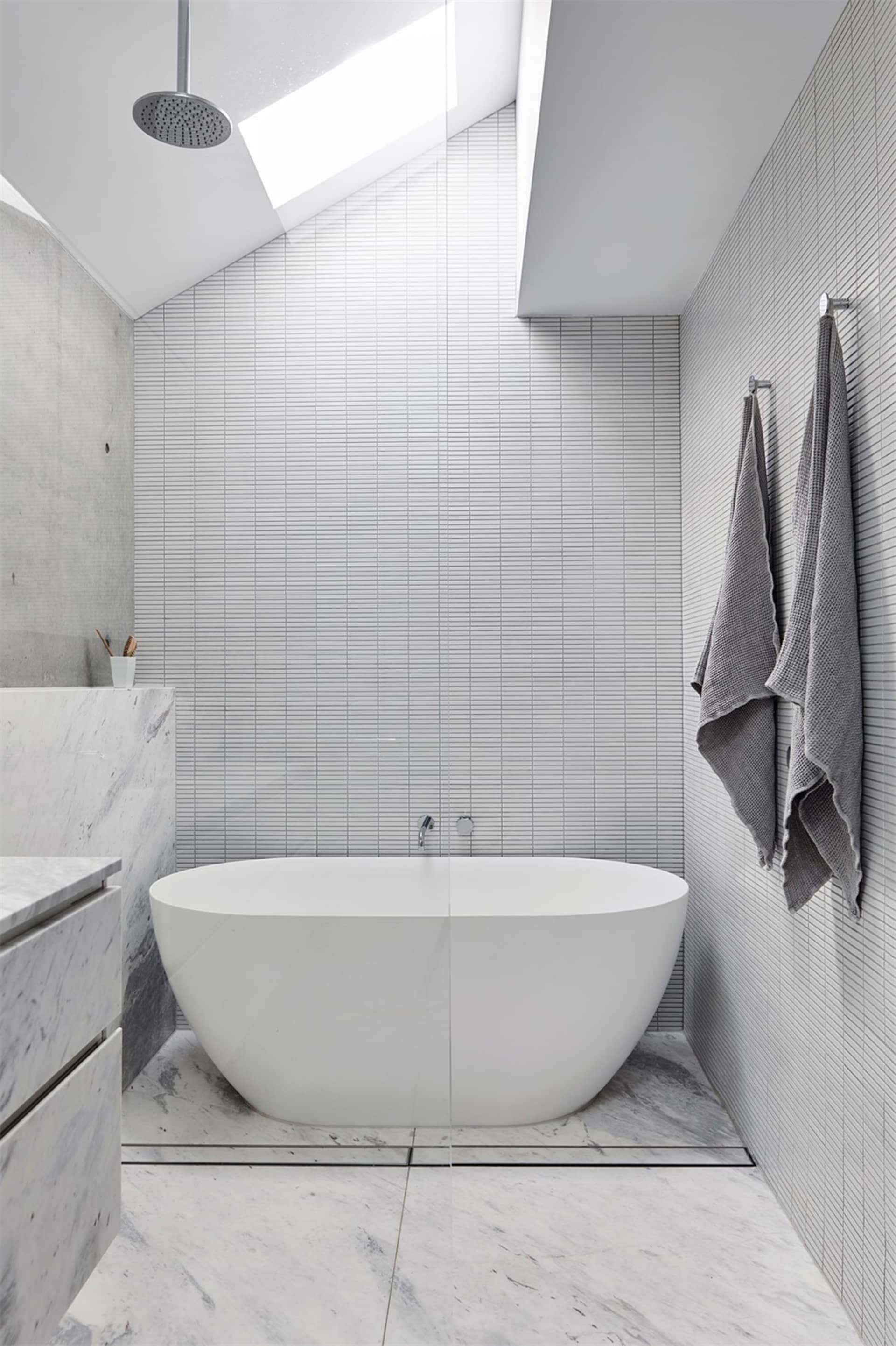 A minimalist bathroom with marble tiles, concrete walls, and a freestanding bathtub under a skylight.
