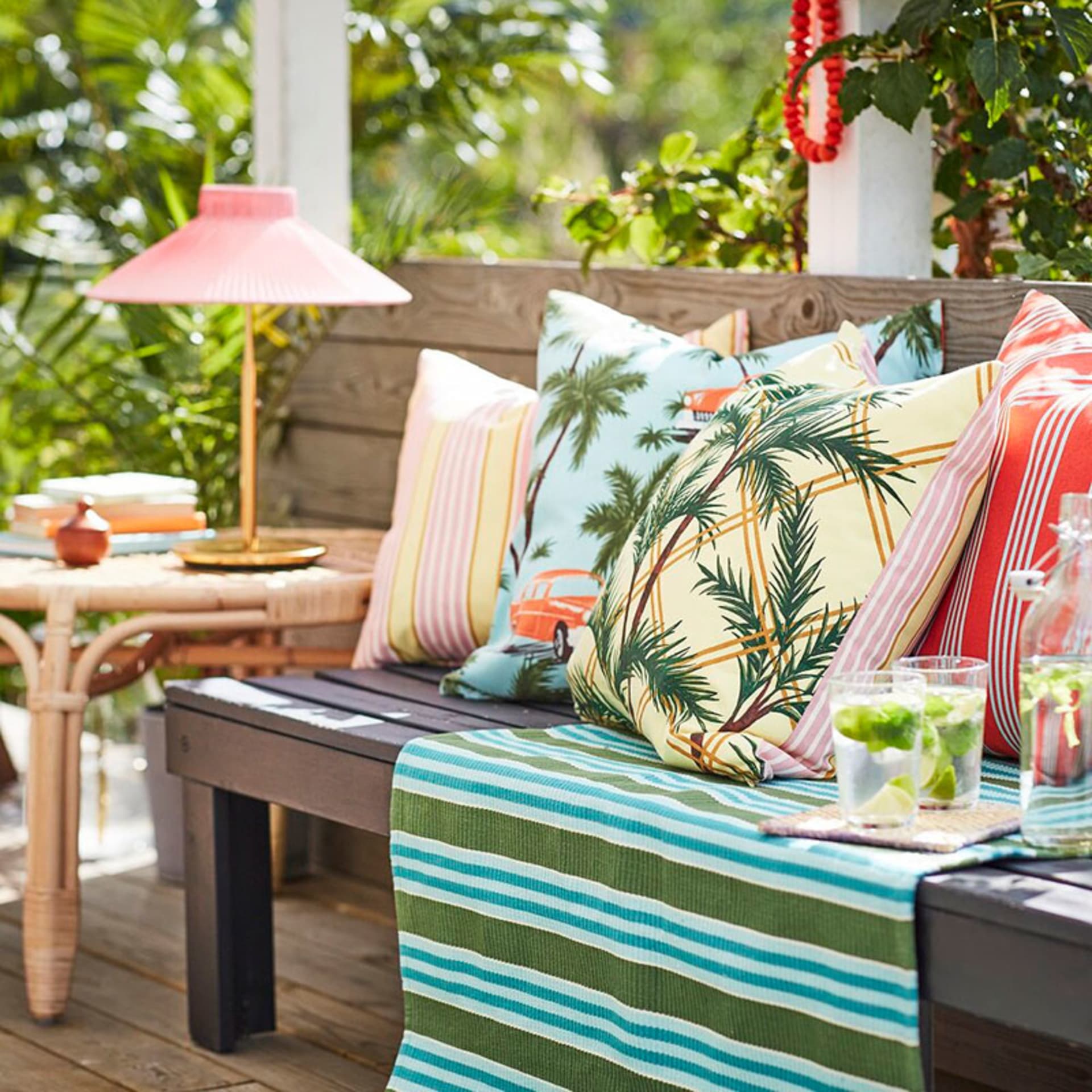 A cozy outdoor seating area with colorful pillows, a striped throw blanket, and a side table with a lamp and drinks.