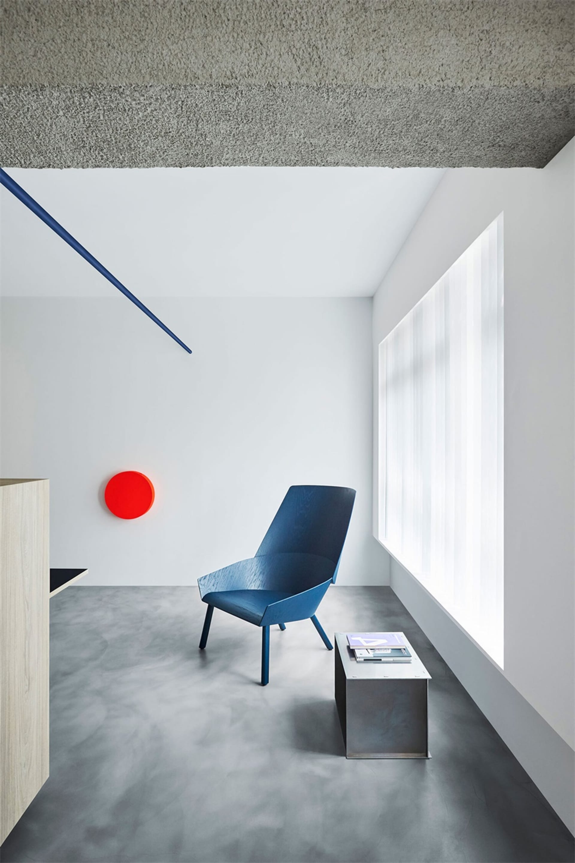 A minimalist living room featuring a blue chair, a red circular light fixture, a concrete floor, and large windows.