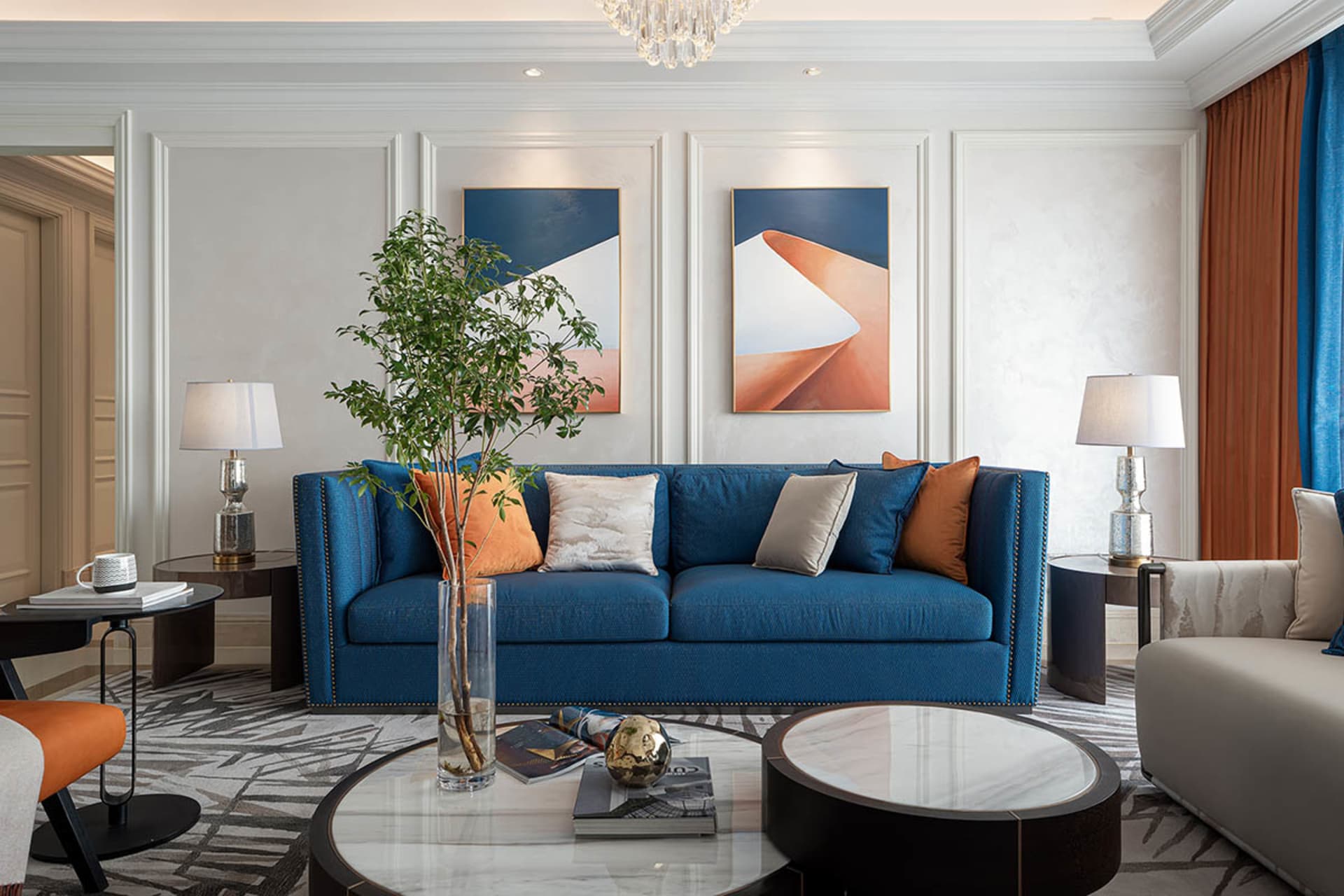 A living room with a blue upholstered sofa, decorative pillows, a round glass coffee table, and framed abstract art on the wa