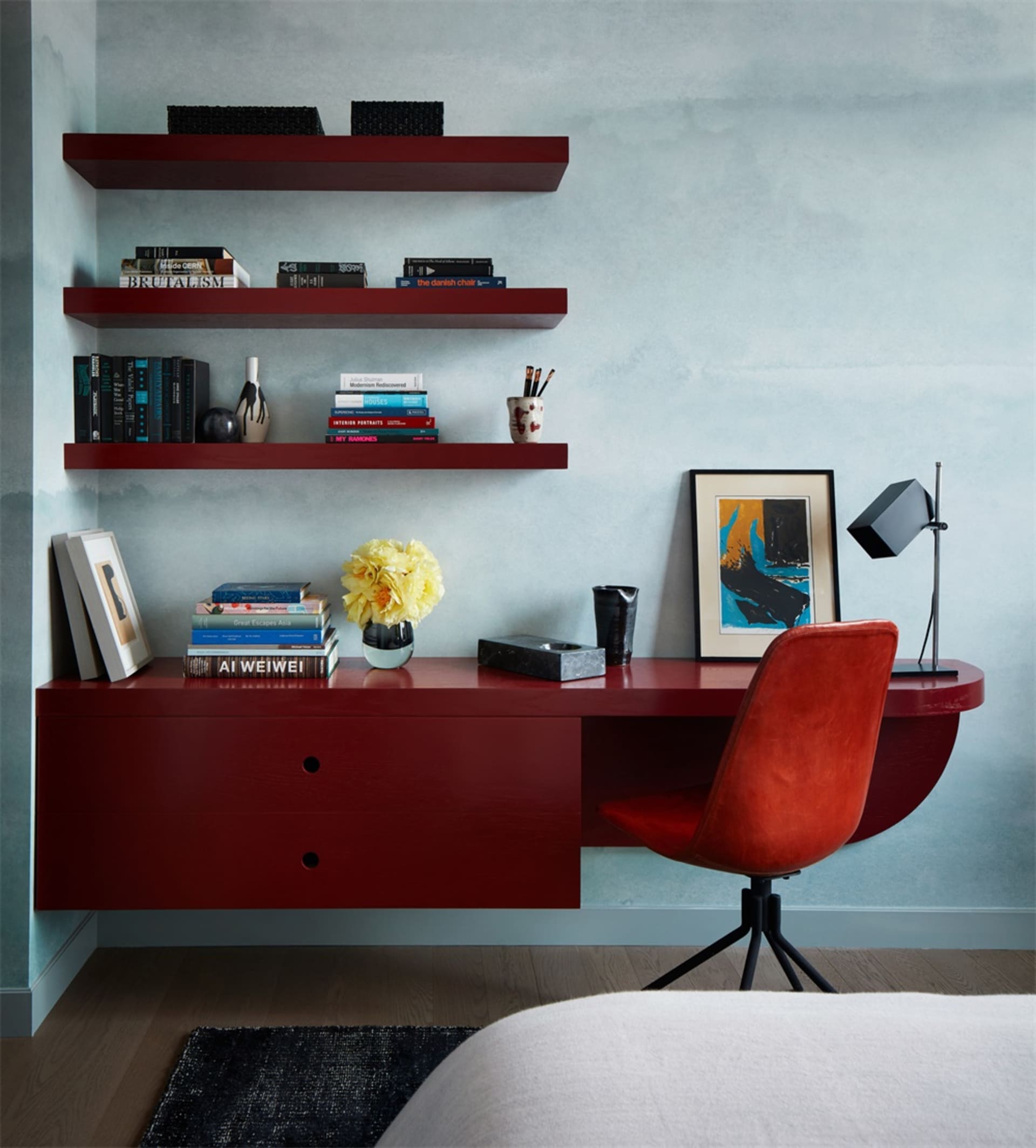A modern home office setup featuring a maroon desk and wall-mounted shelves with books, a vase of flowers, and other decorati
