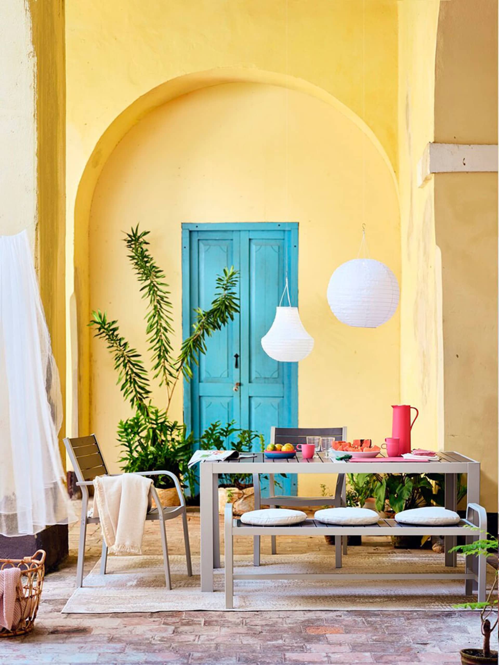 Outdoor dining area with a wooden table, chairs, and hanging lanterns against a yellow and blue arched wall.