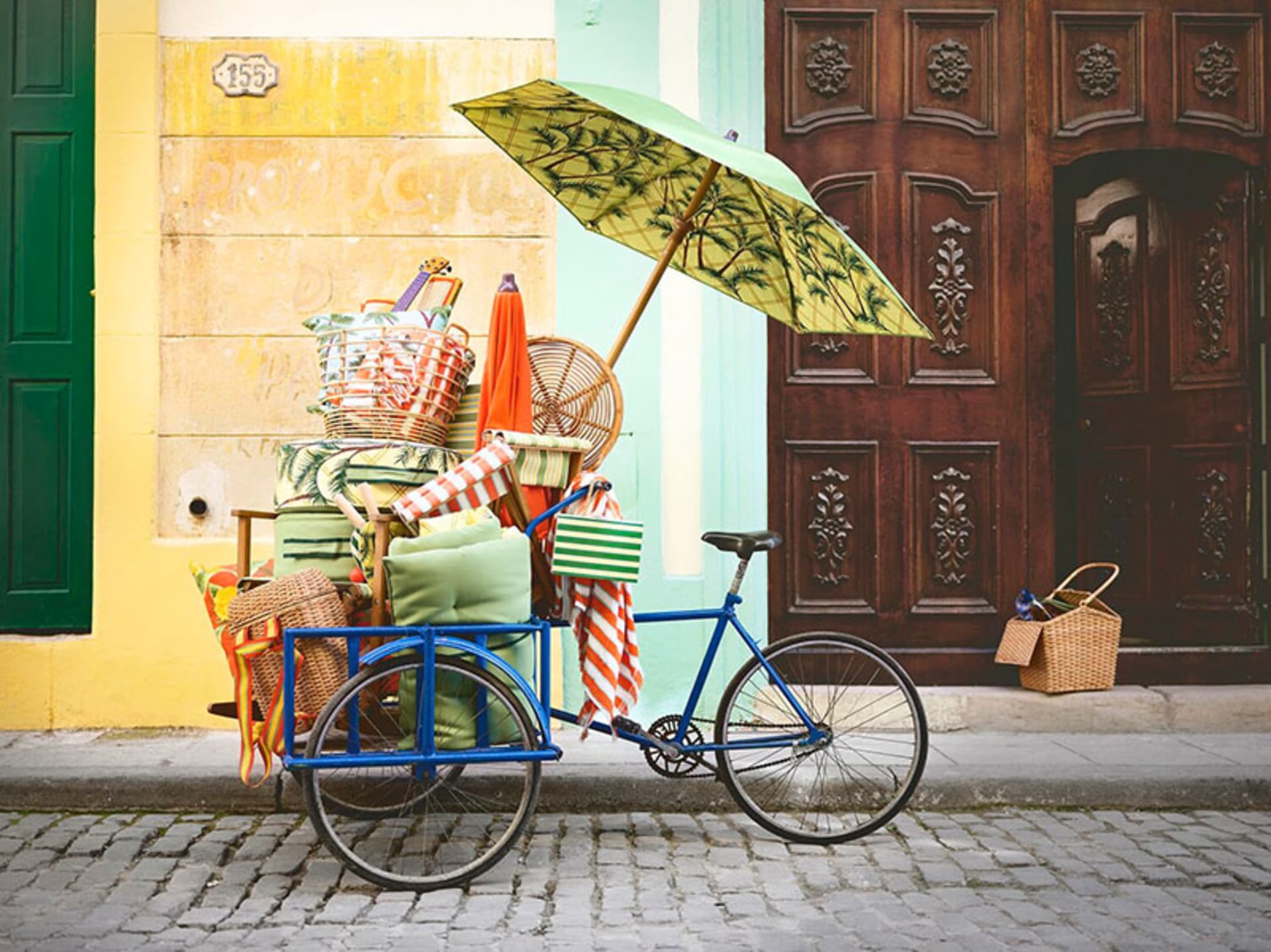 A blue bicycle cart laden with picnic baskets and a green bamboo-patterned umbrella on a cobblestone street next to a wooden