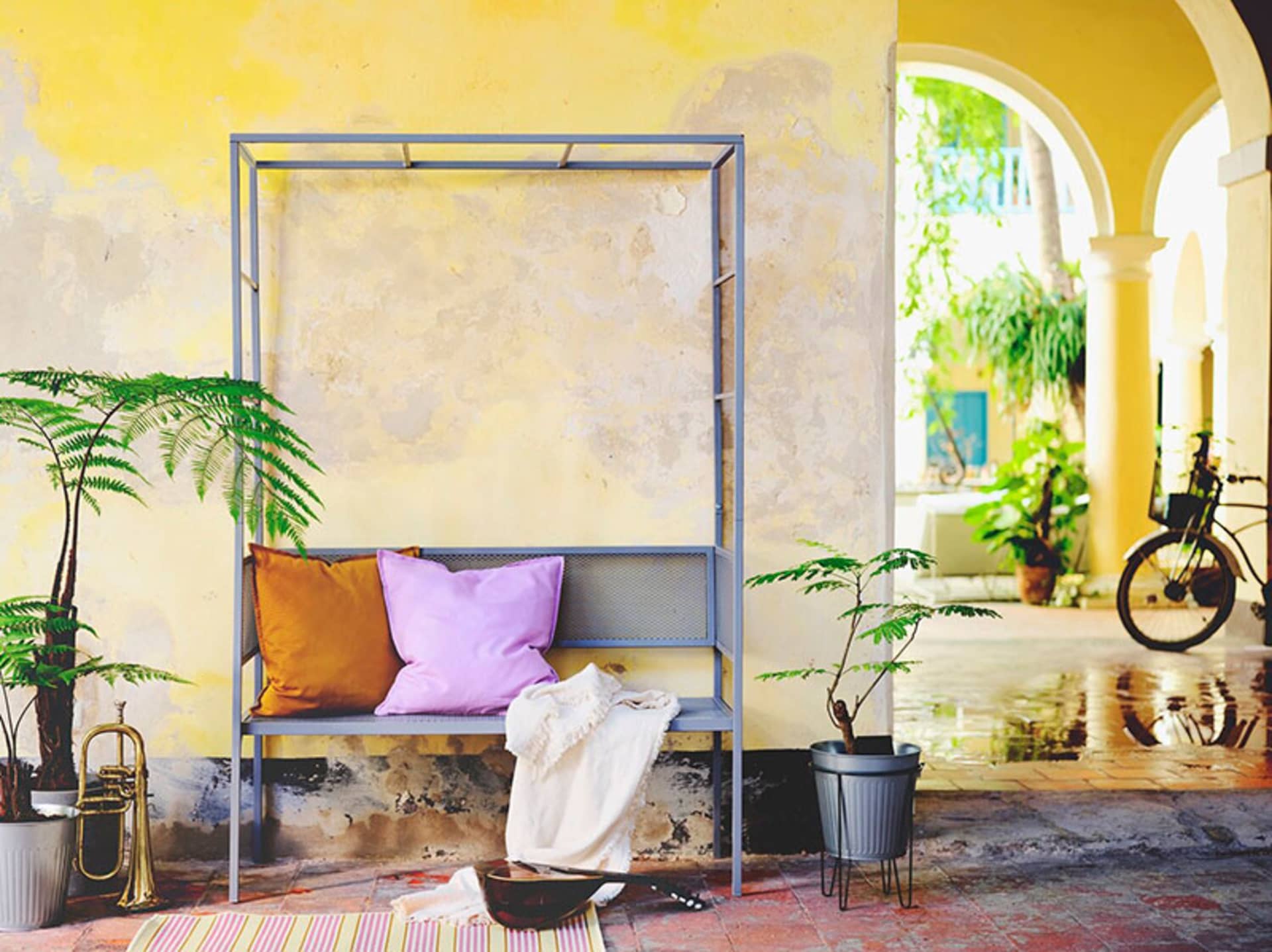 A modern interior decor image featuring a metal frame with purple and mustard pillows on a bench, surrounded by potted plants