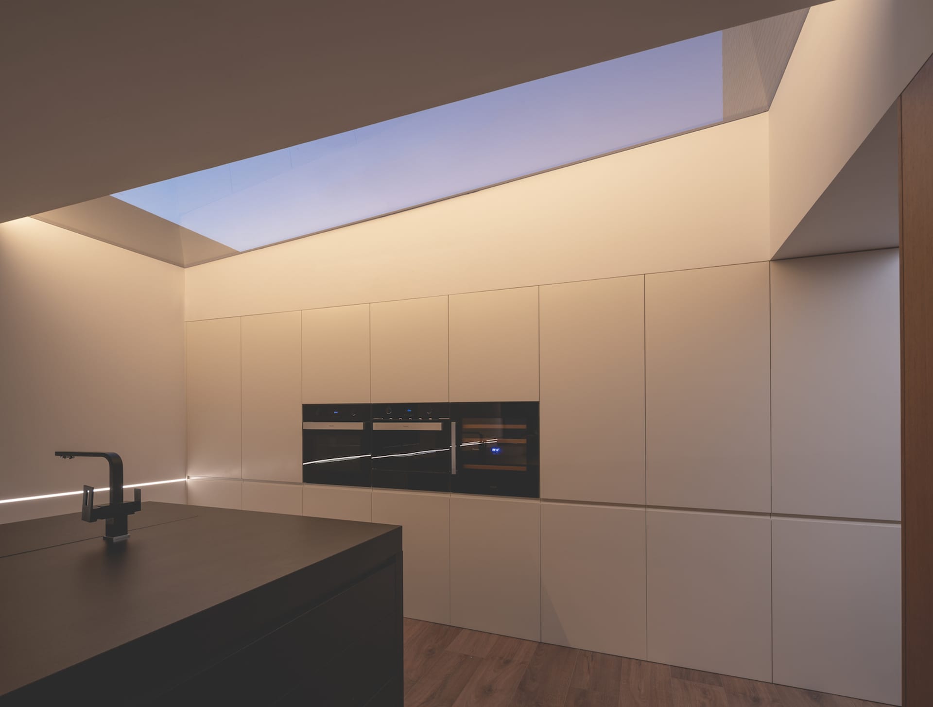 Modern kitchen featuring minimalist design with white cabinets, wood flooring, and a glass skylight.