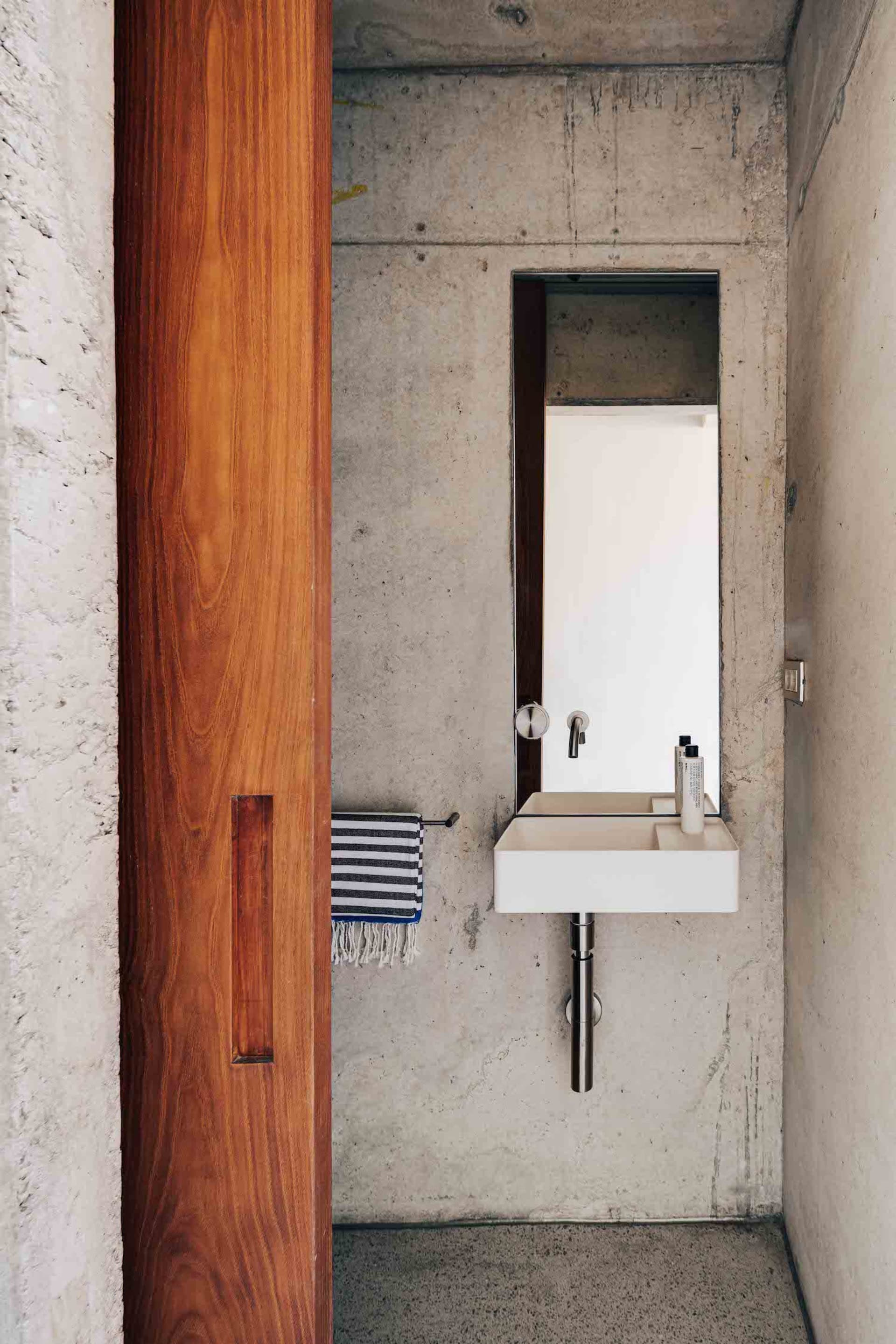 A minimalist bathroom with concrete walls, a wooden door, a white sink, and a striped hand towel hanging on a towel ring.