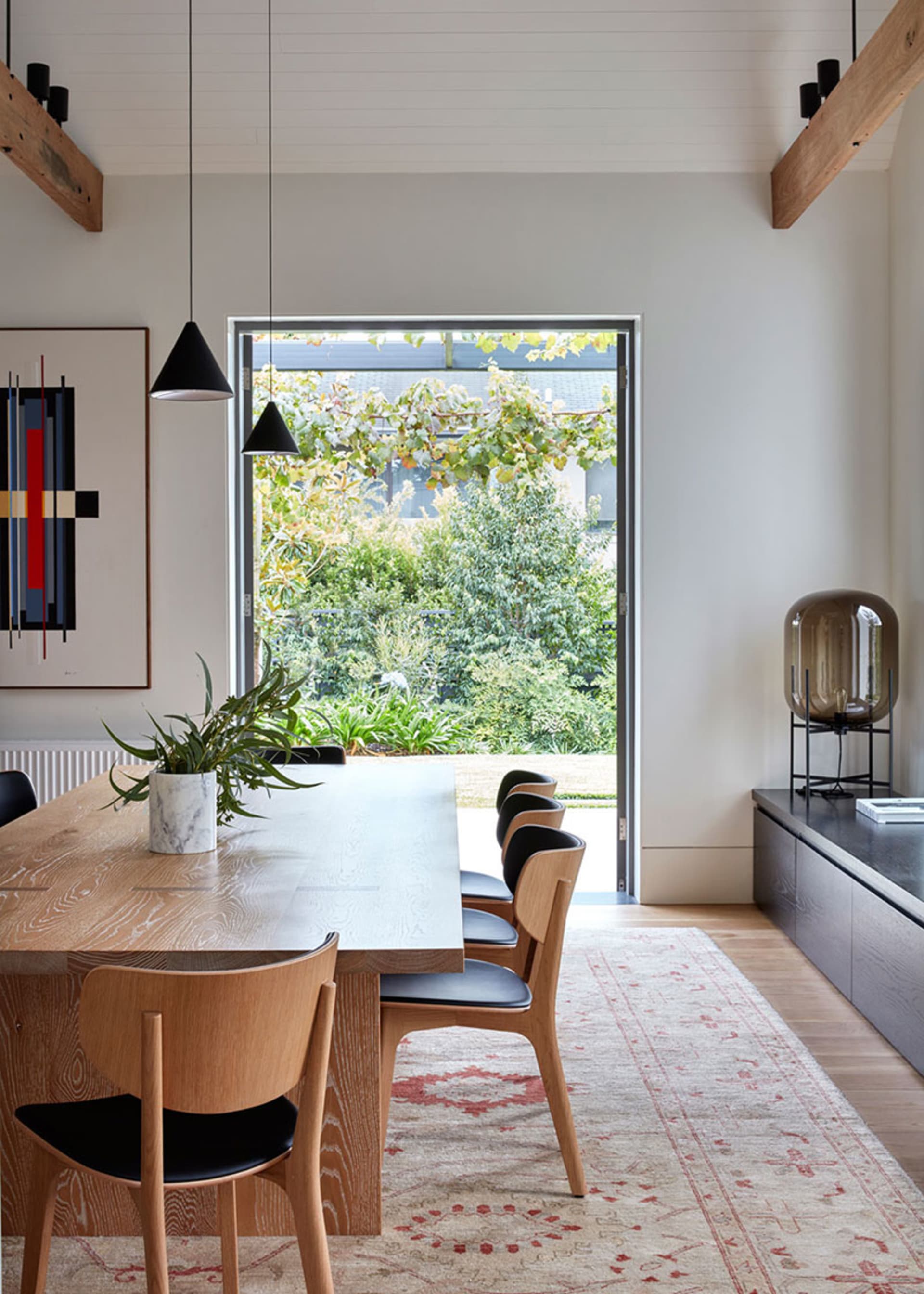 A contemporary dining room featuring a wooden table with six chairs, a patterned rug, and a large window overlooking greenery