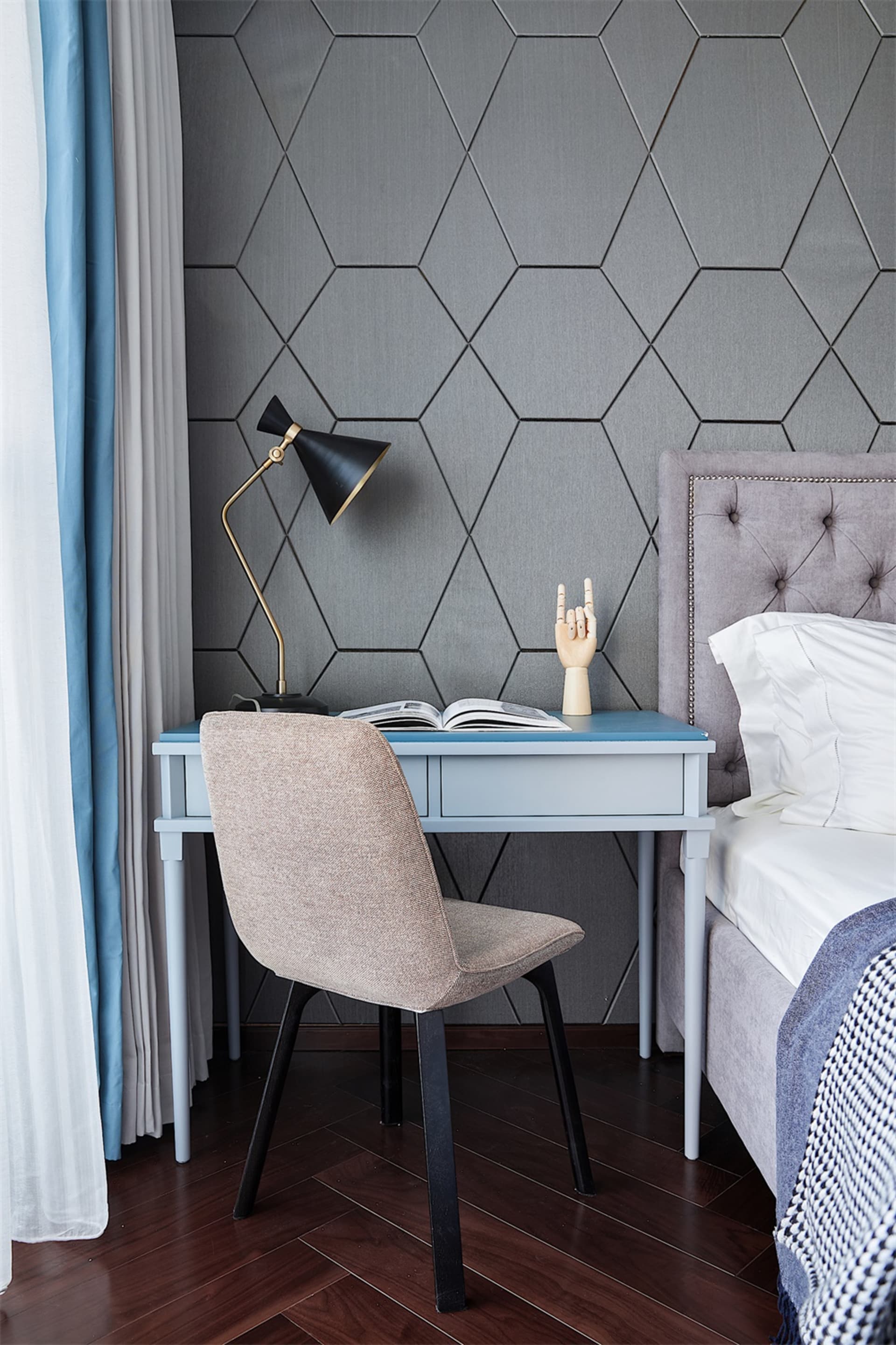 A minimalist bedroom featuring a workspace with a blue desk, beige chair, black table lamp, and a hexagonal patterned wall, a