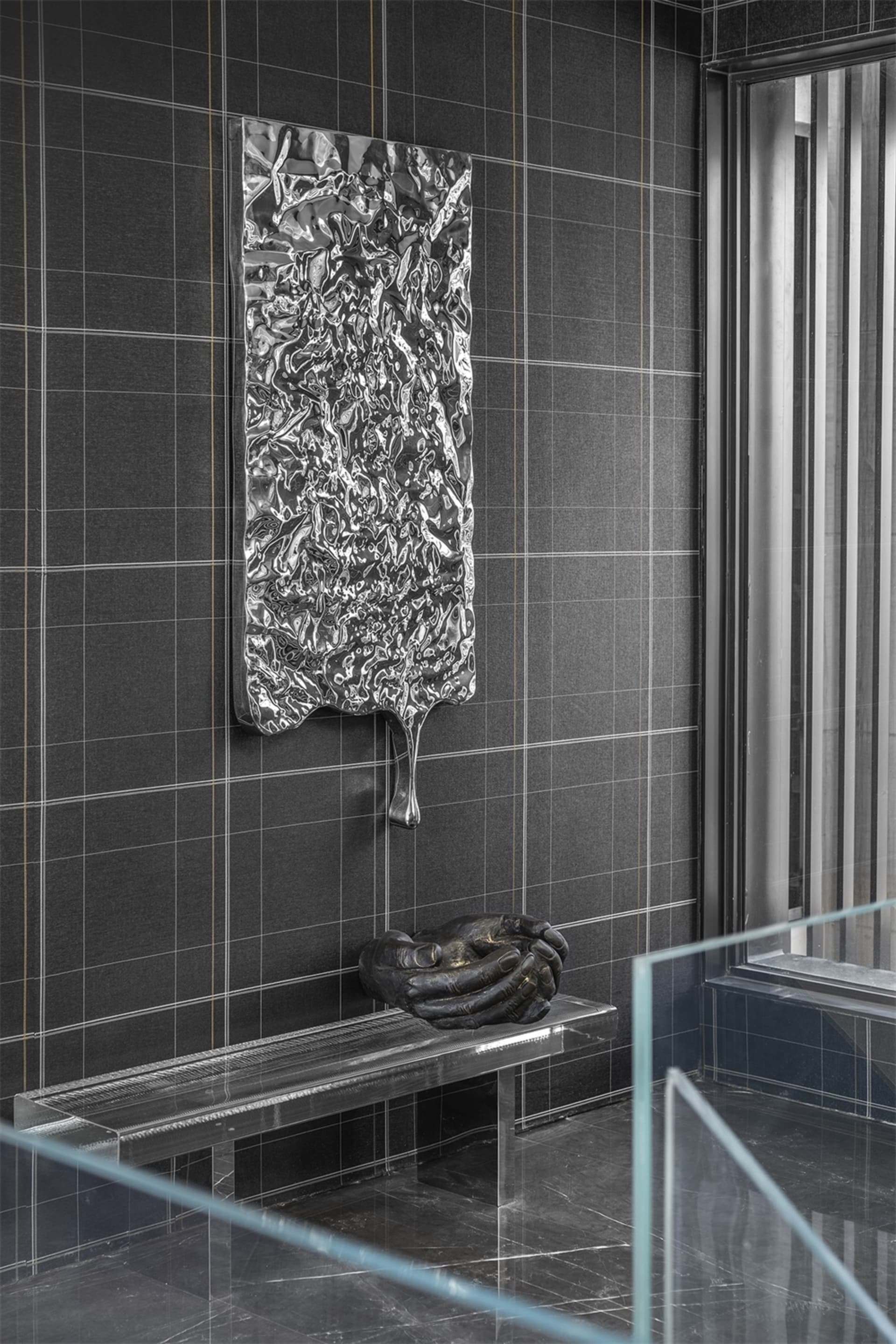 A metallic panel with a crumpled surface texture is mounted on a dark grid-tiled wall, accompanied by a sculptural hand gestu