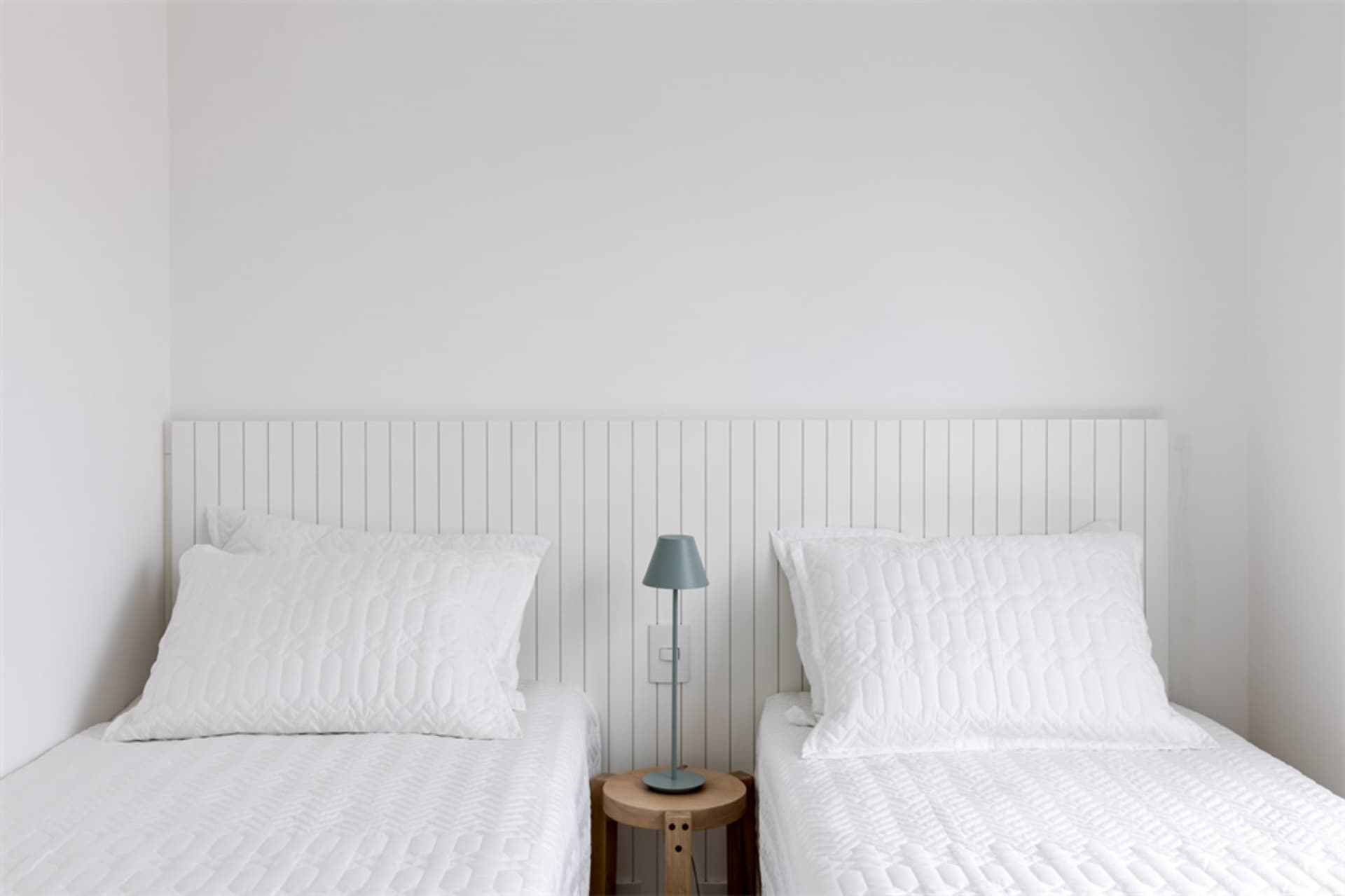 A minimalist bedroom with two beds, white bedding, and a small wooden table with a lamp.