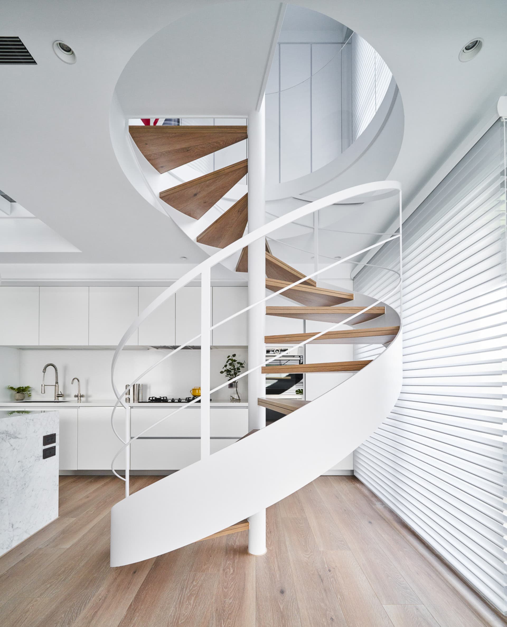 A modern, minimalist kitchen featuring a spiral staircase with white metal railings and wooden steps, designed with sleek, fu