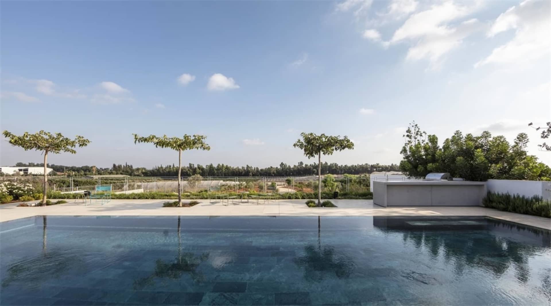 A modern exterior garden with a blue tiled pool, concrete tiles, and a view of distant trees and sky.