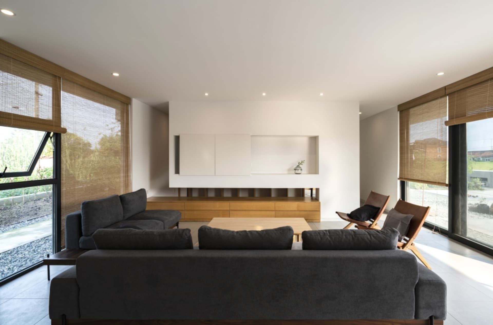 Modern minimalist living room with dark gray sofas, wooden coffee table, and glass vase on wooden shelf, framed by large wind