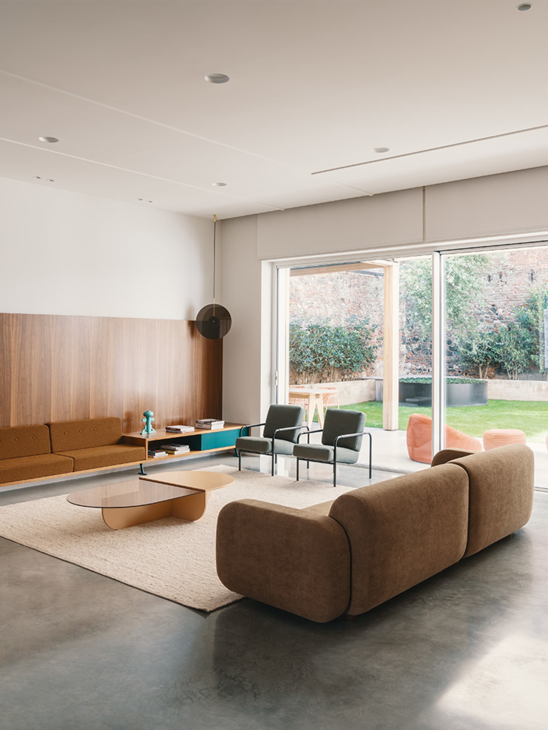 A modern living room with concrete flooring, a brown sectional sofa, a round glass coffee table, and a wooden shelf with deco