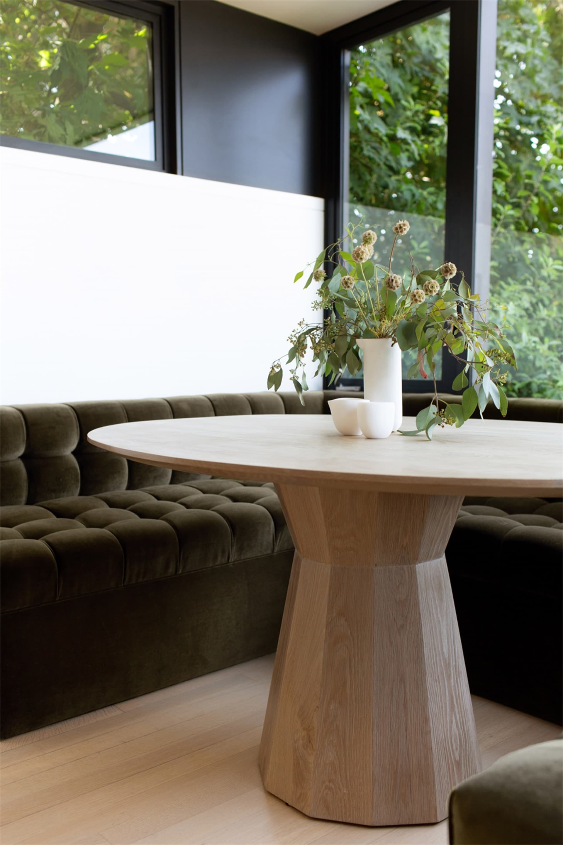 A modern living room featuring a wooden table adorned with greenery and two white cups, situated in a corner with large windo