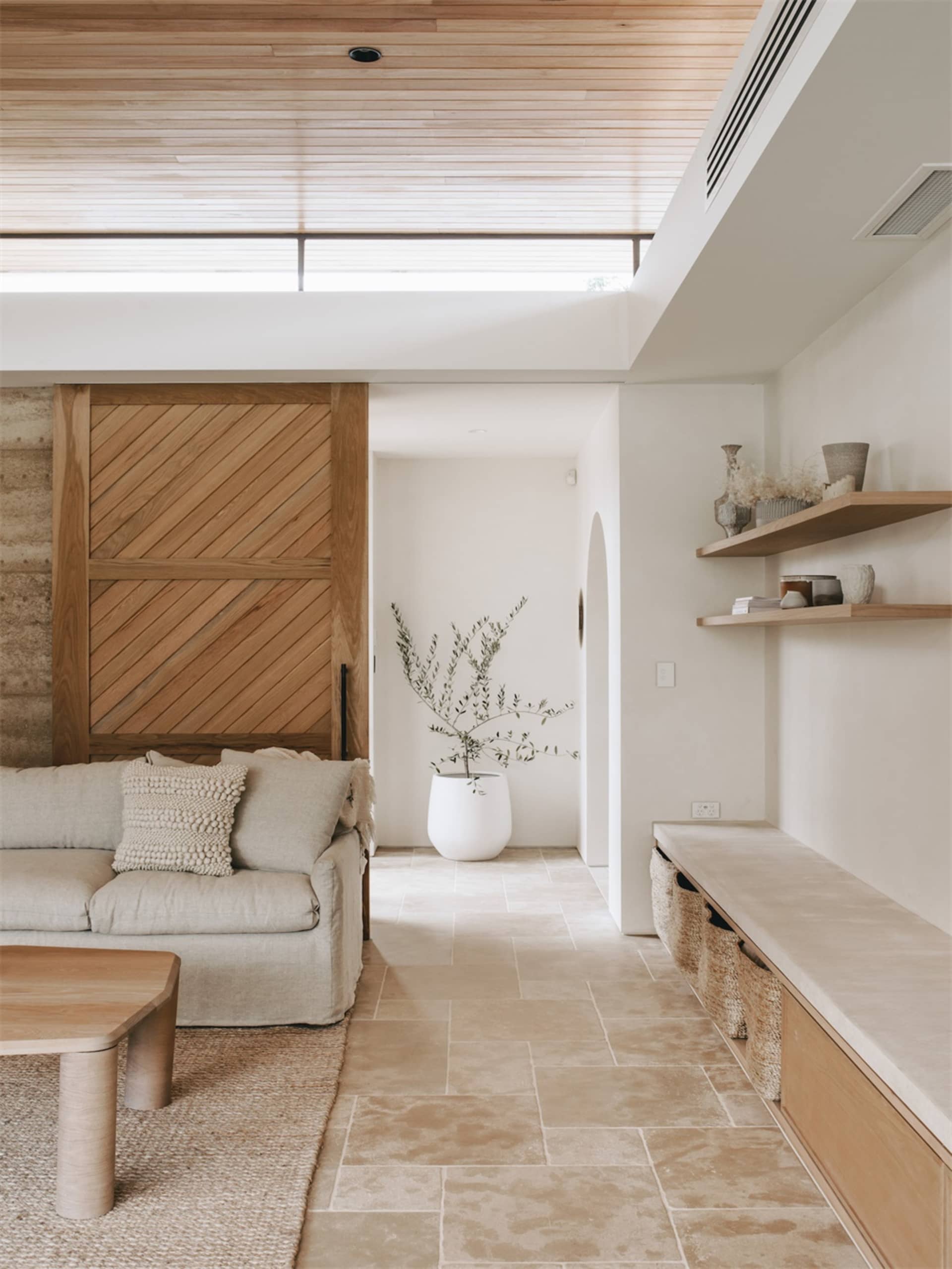 A minimalist living room featuring a beige couch, a wooden table, and a concrete floor. The room has a sliding wooden door an
