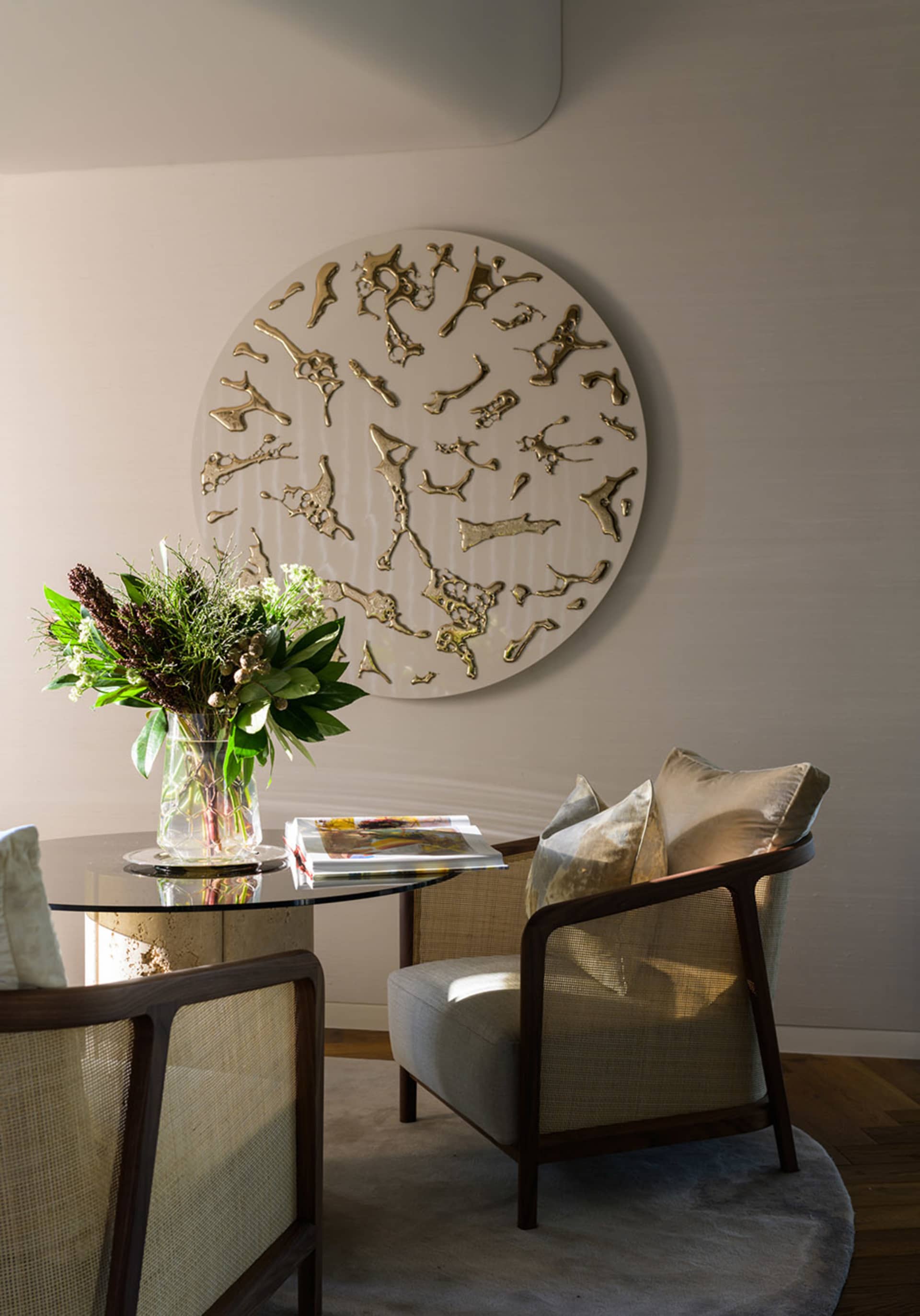 Modern living room with wood and glass elements, featuring a round metallic wall art piece, a glass table, and upholstered ch