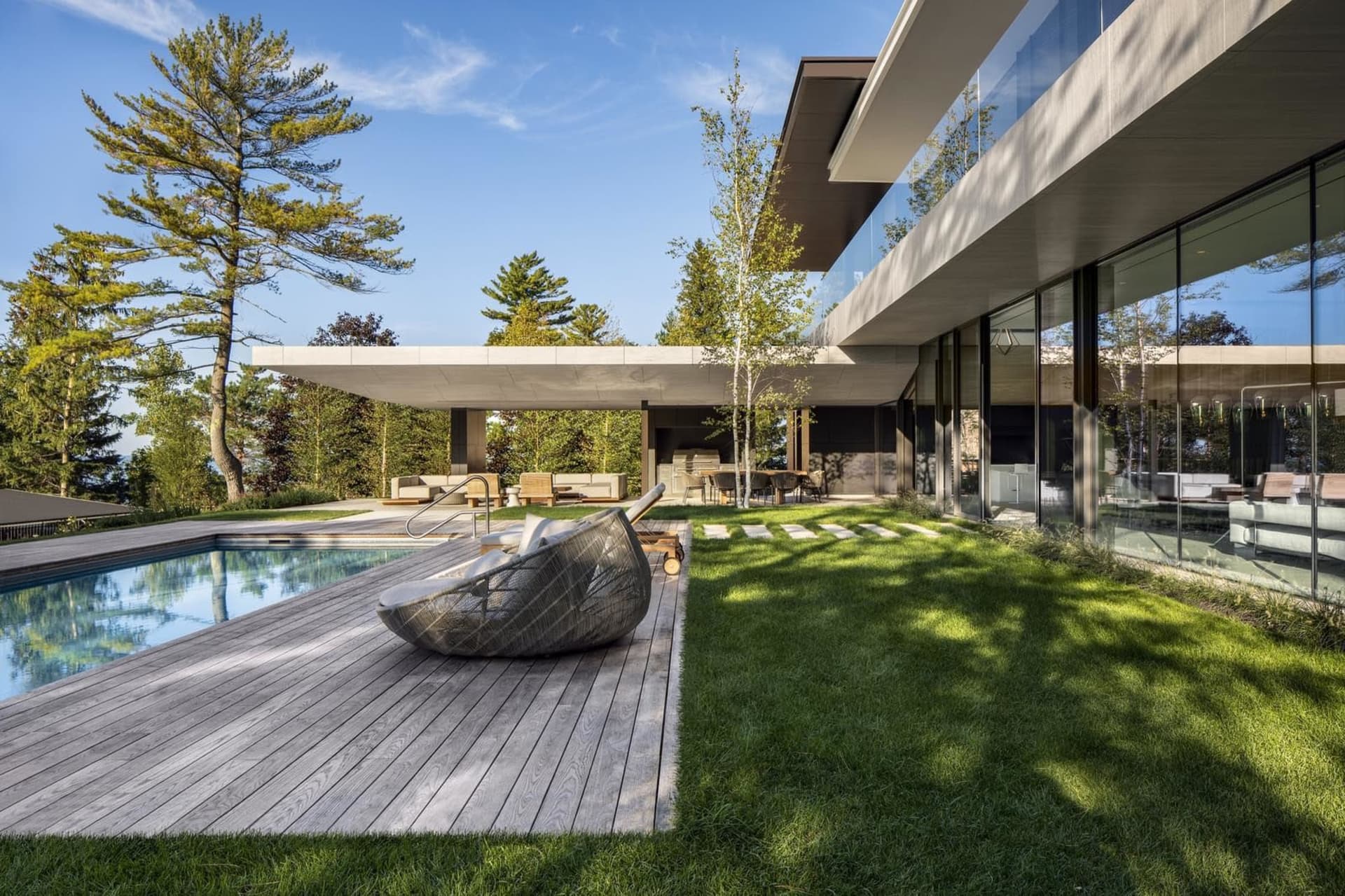 A modern residential exterior featuring concrete and wooden structures, surrounded by greenery and a swimming pool.