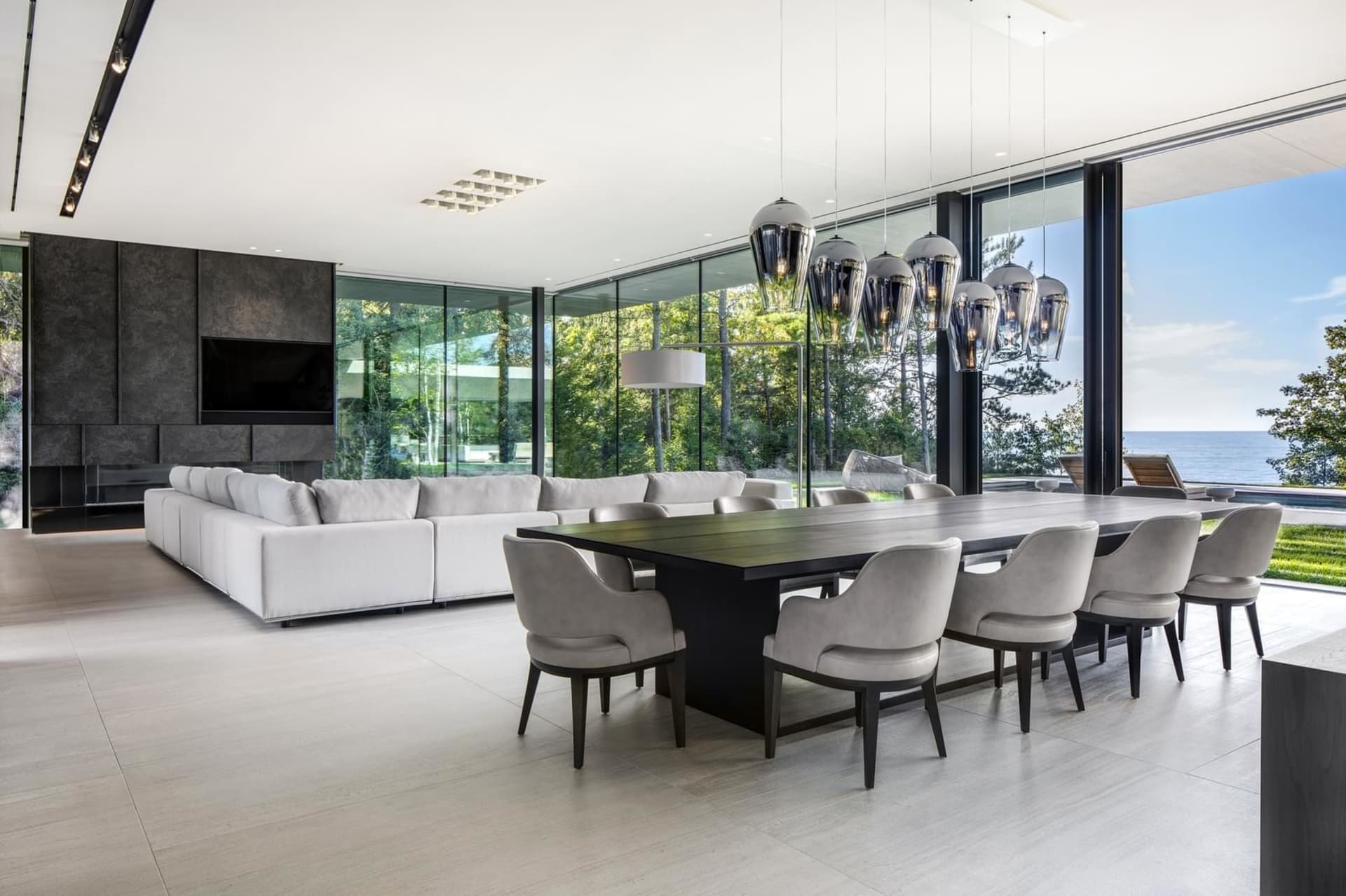 A modern living and dining room with white furniture, wooden table, and large windows overlooking the sea.