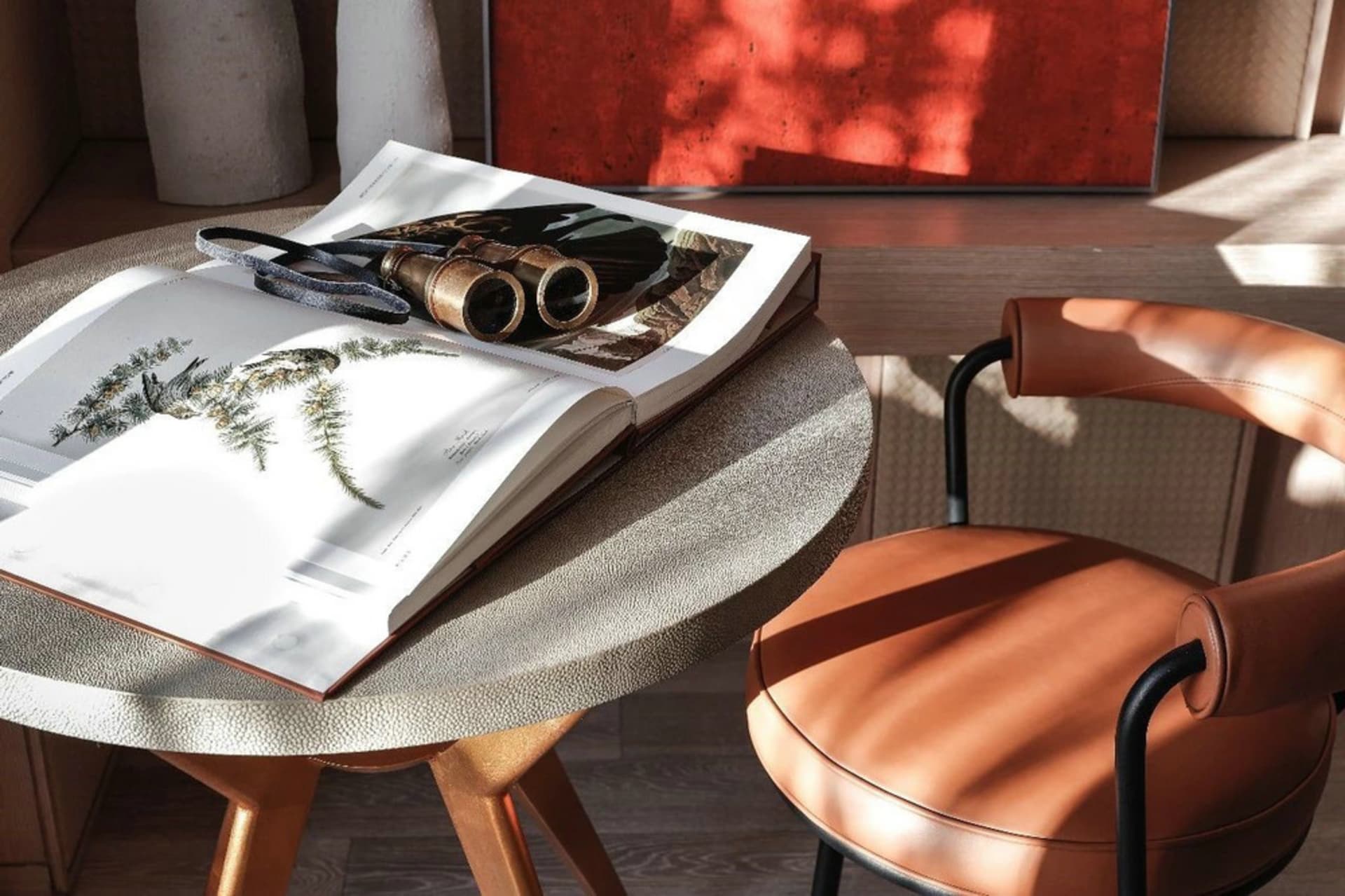 A modern living room featuring a round table with a book, binoculars, and a vase, and a brown leather chair with a cushion an