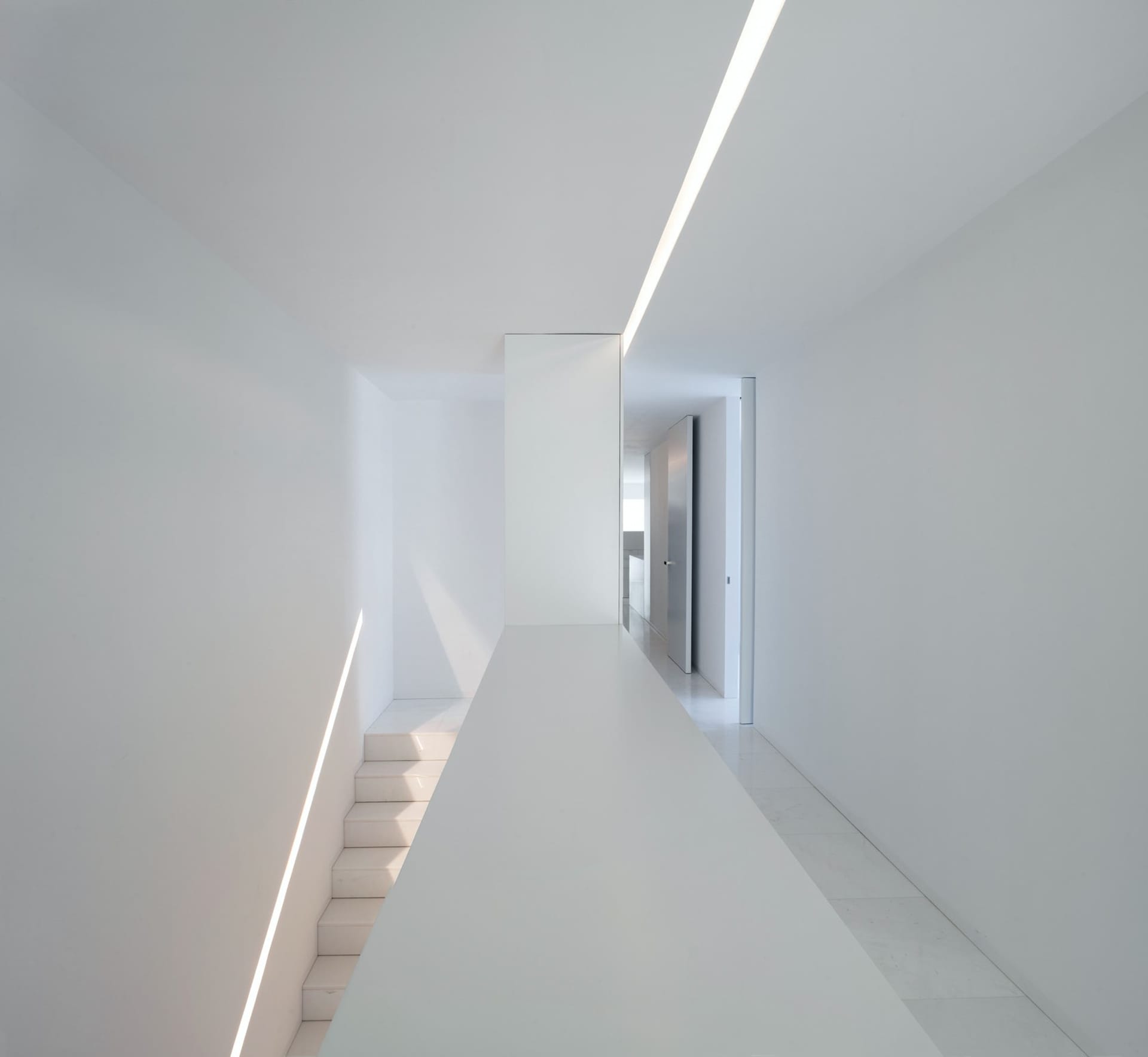A minimalist white interior with a white wall and floor, featuring a staircase with white steps and a light source from above