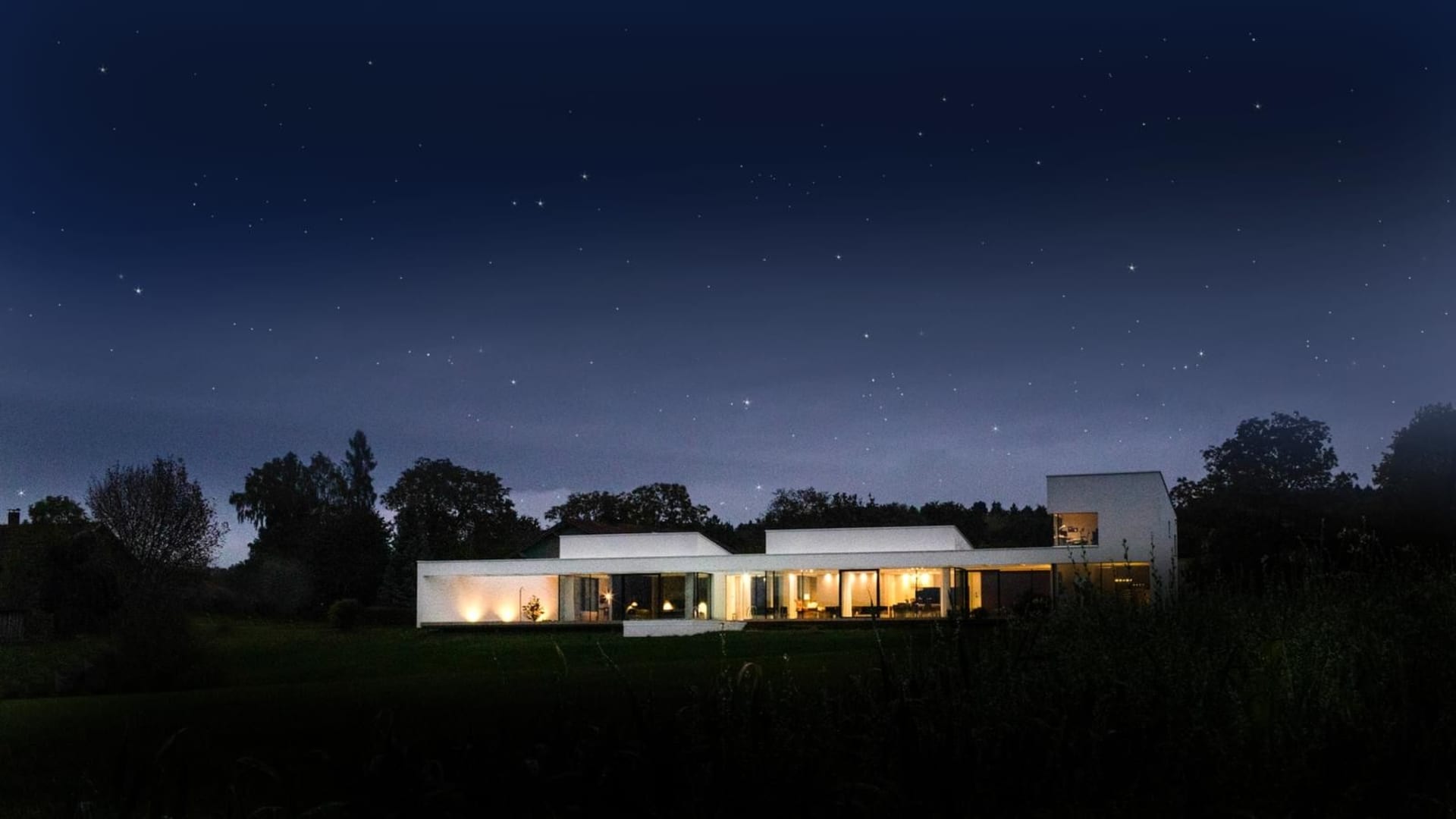 A modern, minimalist residential building with large glass windows and a white exterior at night with a starry sky.