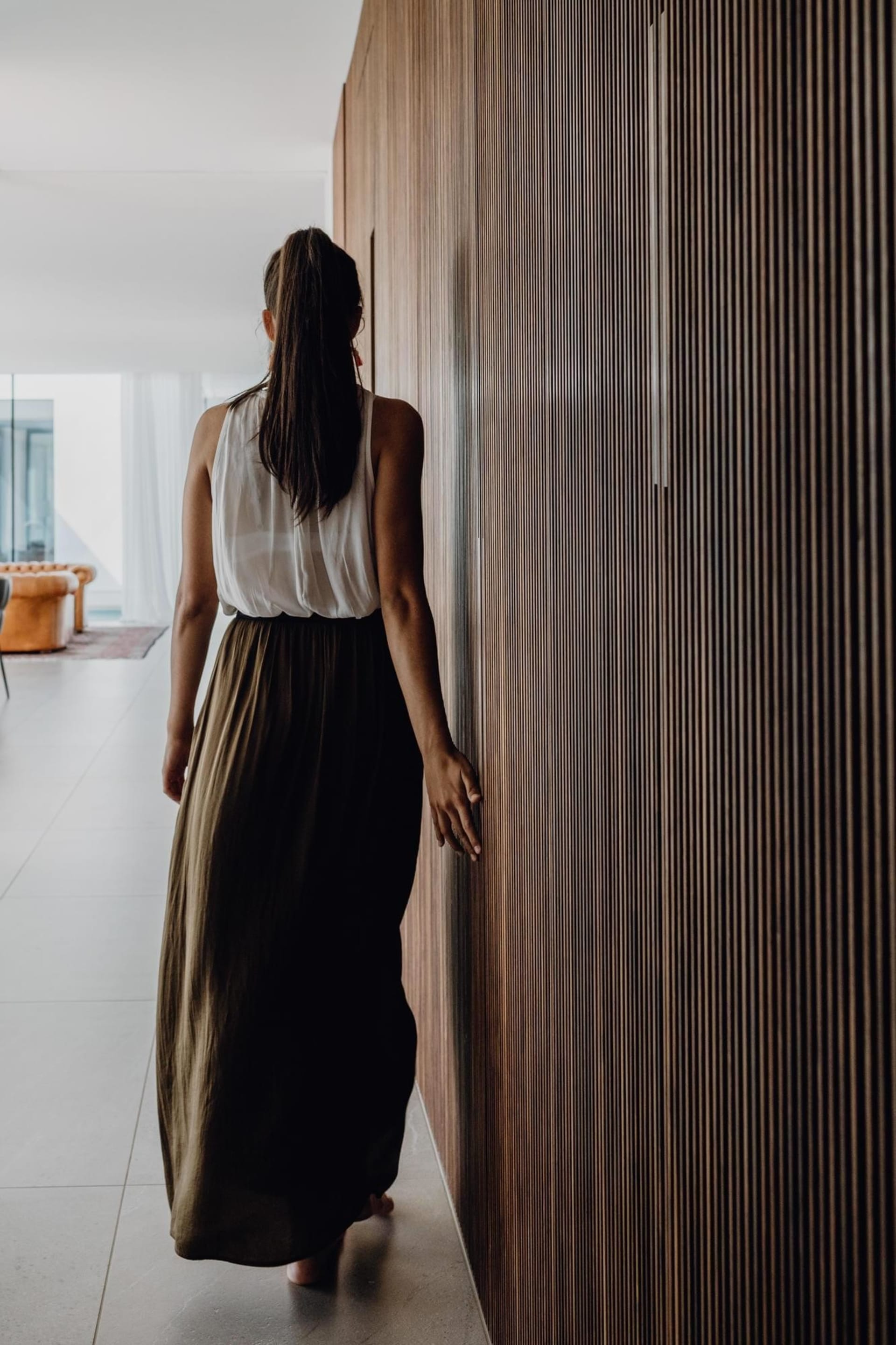 a woman wearing a white shirt and black skirt walking in front of a tall, striped wall, possibly in a modern or minimalist in