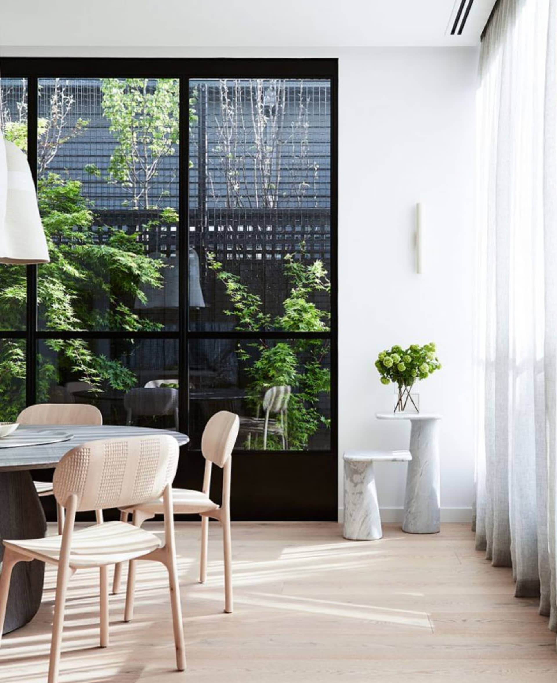 A minimalist dining room featuring wooden chairs and a marble table, surrounded by large windows.